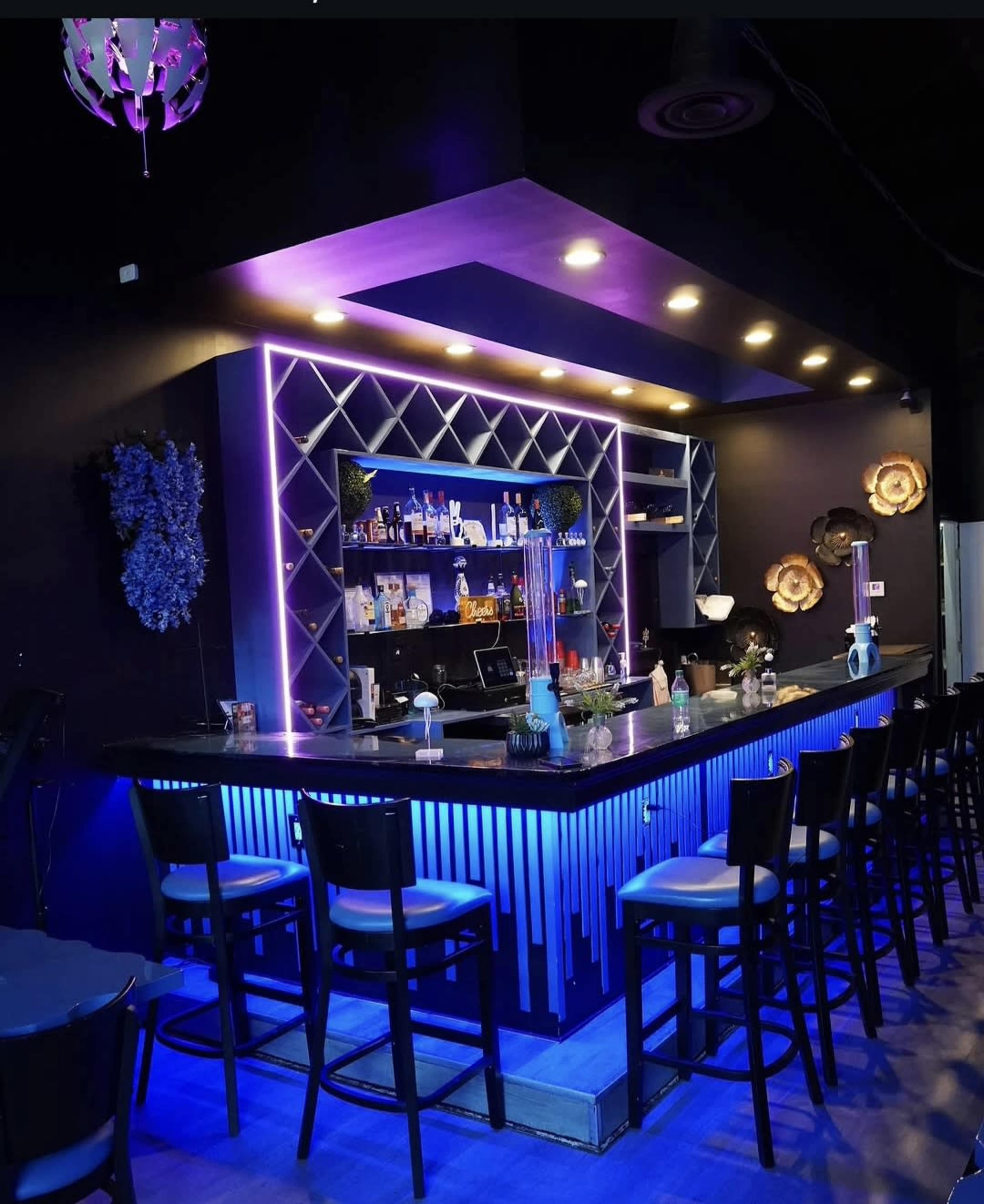 The image shows a modern bar area with a backlit shelf displaying various bottles, illuminated by purple and blue lighting, and featuring a long counter with black chairs.