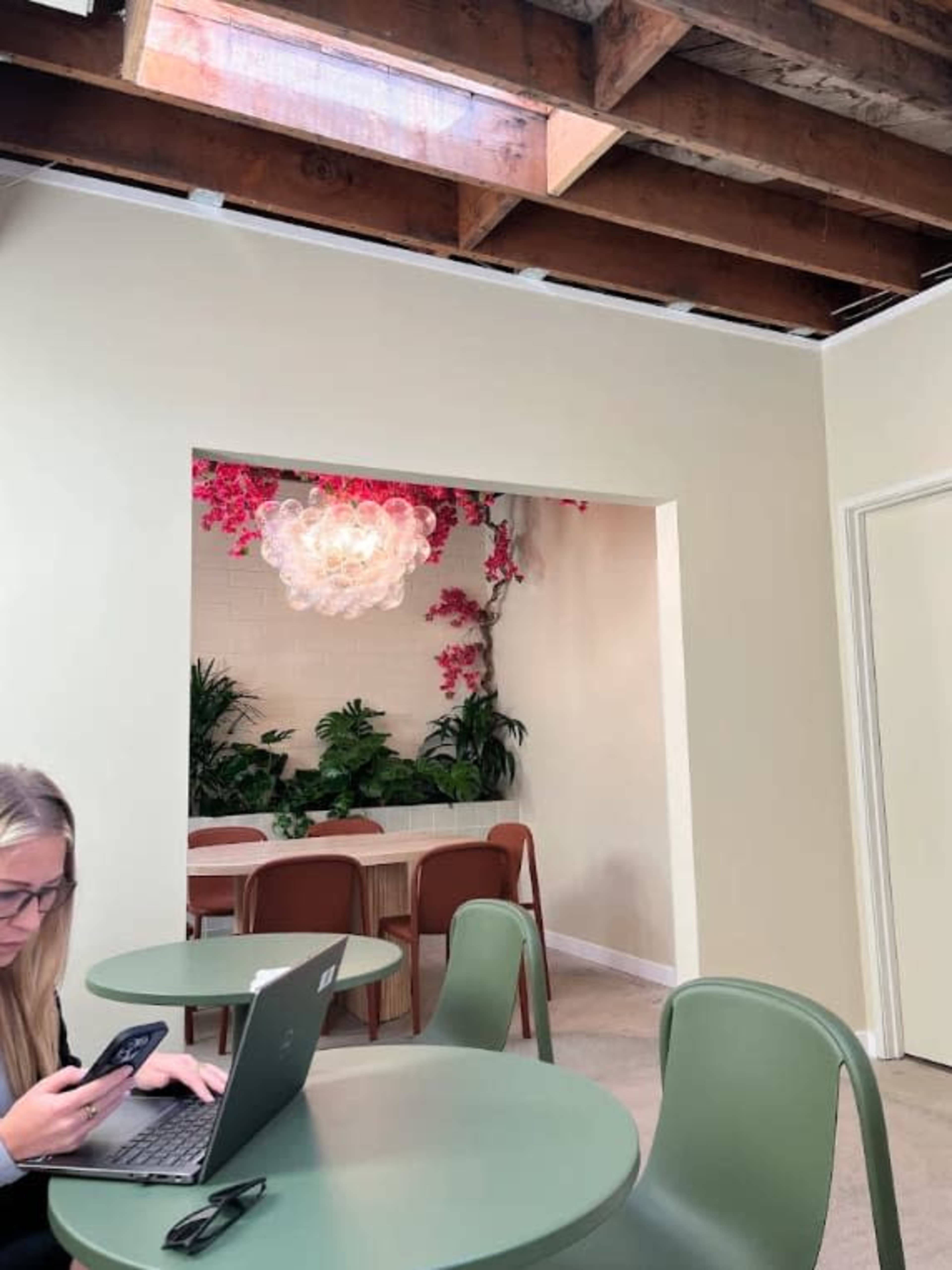A woman sits at a green table using a laptop and phone, while a bright area with a floral chandelier and greenery is visible in the background.