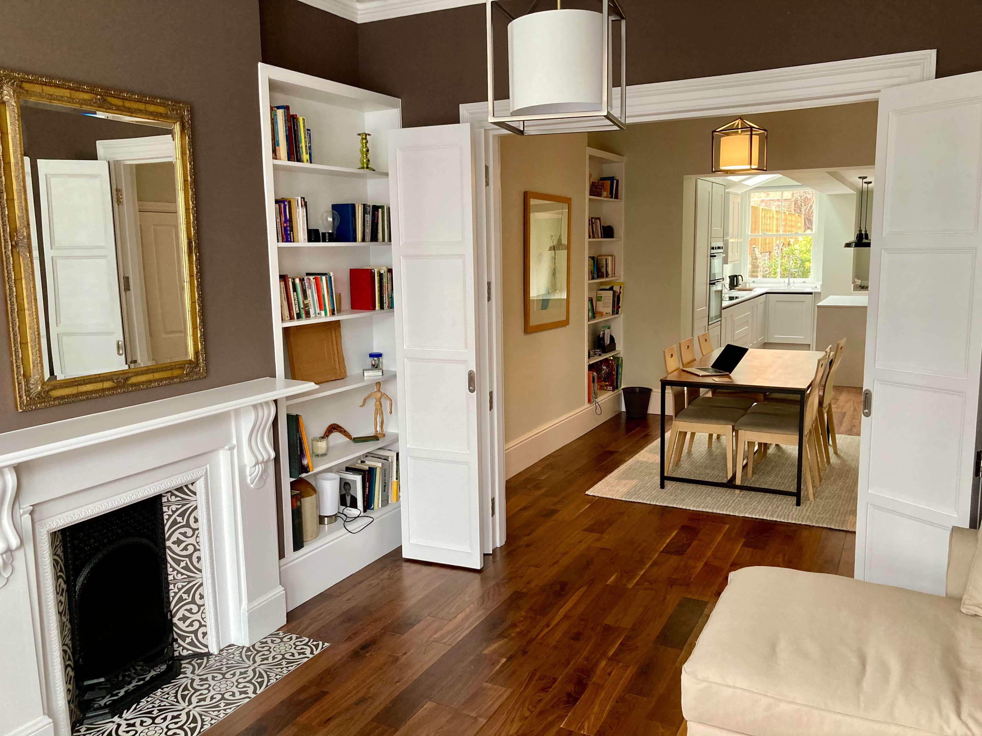 The image shows a cozy interior with a living room featuring a fireplace and bookshelves, leading to a dining area with a long table and open kitchen.