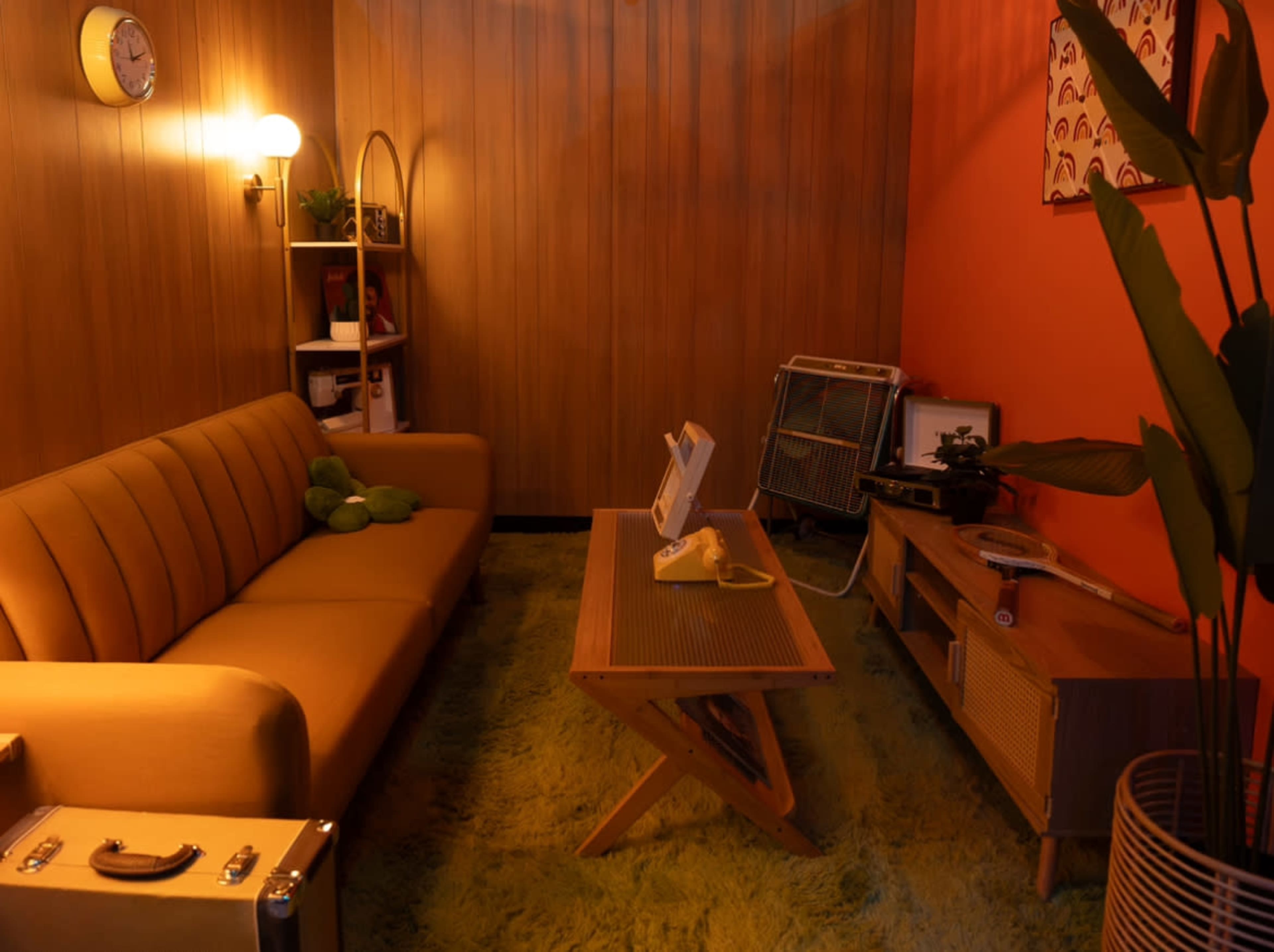 A retro-inspired living room with a yellow sofa, a wooden coffee table, a vintage telephone, and shelves filled with decorative items against a backdrop of wood paneling and an orange wall.