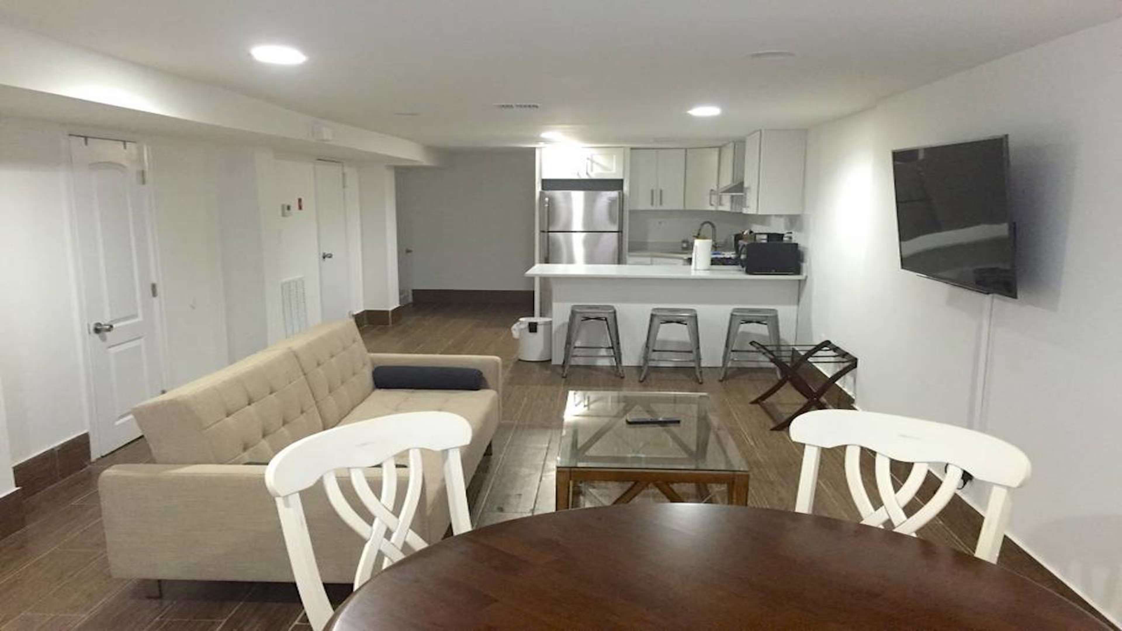 The image shows a modern, open-concept living space featuring a kitchen with stainless steel appliances, a beige sofa, a glass coffee table, a dining table with white chairs, and a mounted television.
