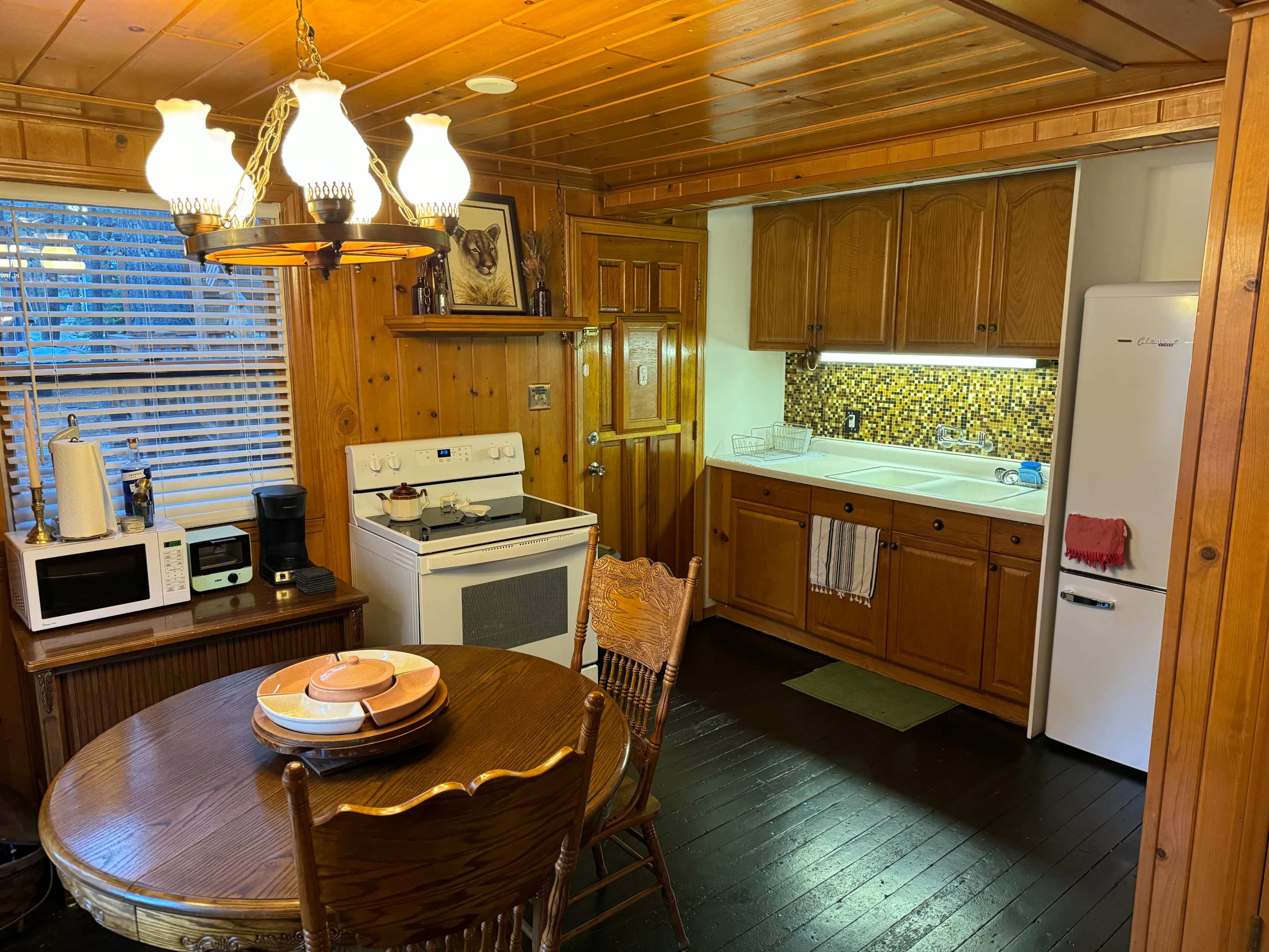 A wooden kitchen features a round table with chairs, an electric stove, a refrigerator, and cabinets with a mosaic backsplash.