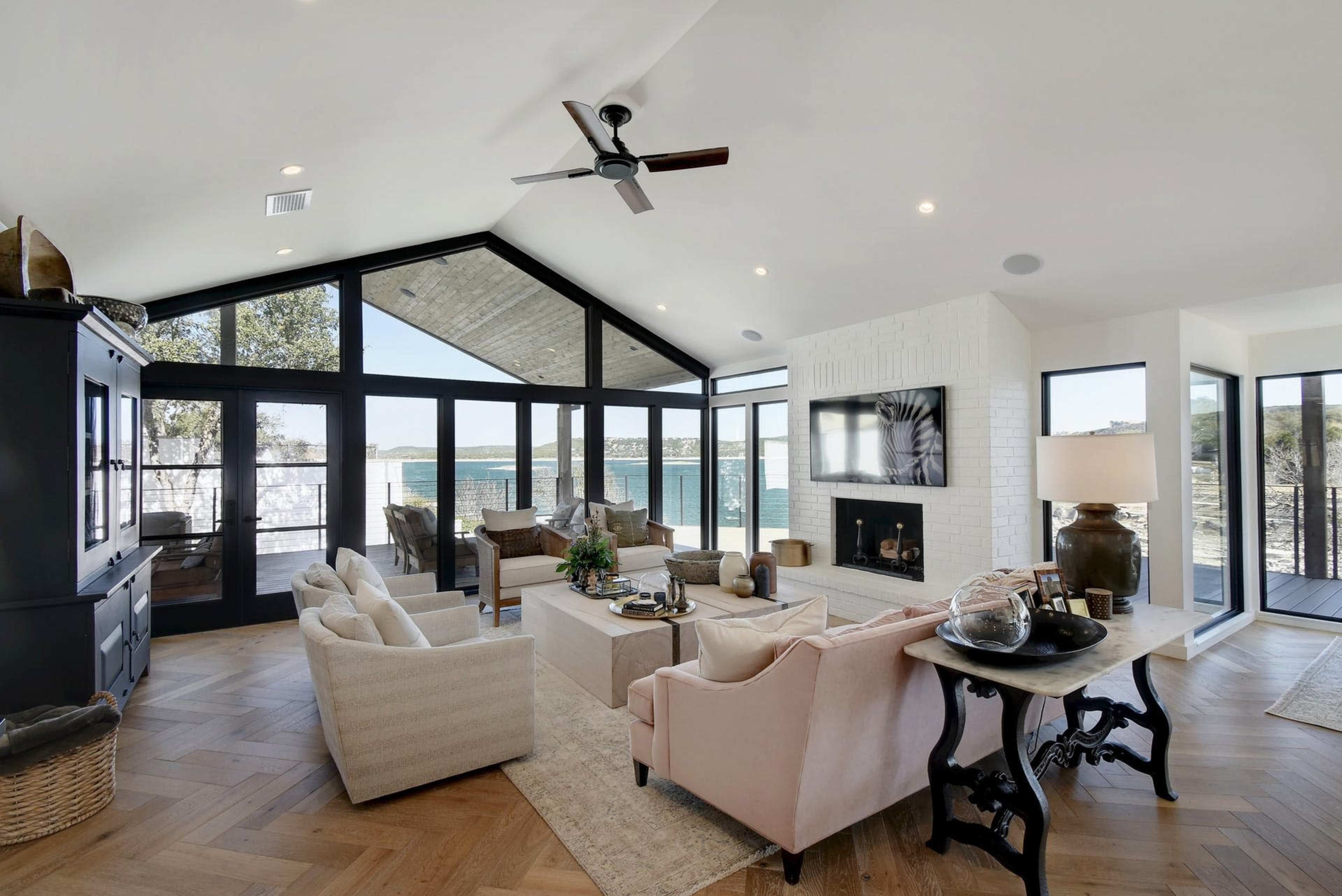 A spacious living room features large windows overlooking a body of water, with a mix of seating arrangements and a modern fireplace.