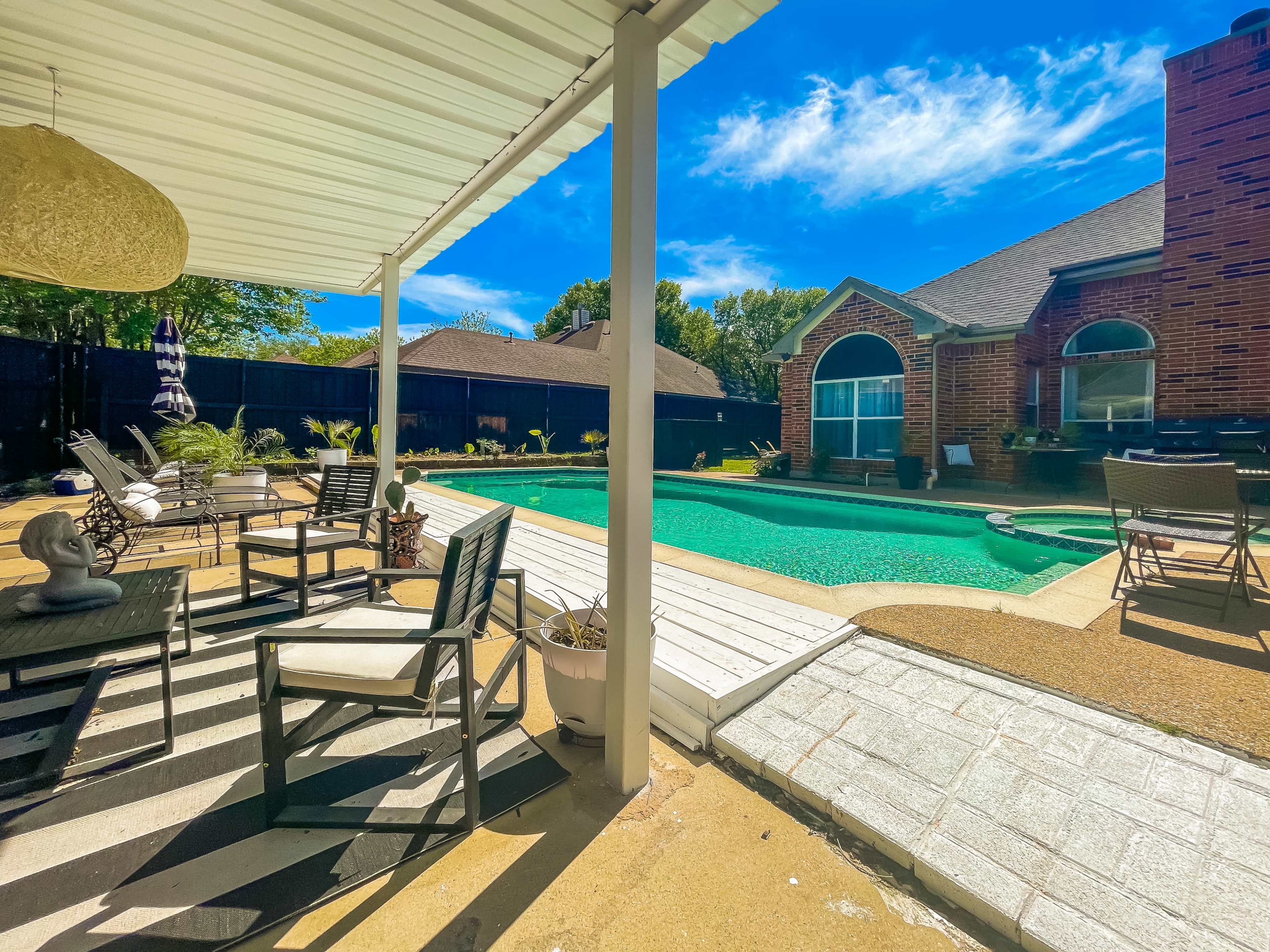 A backyard with a swimming pool surrounded by patio furniture and a covered area with a light fixture.