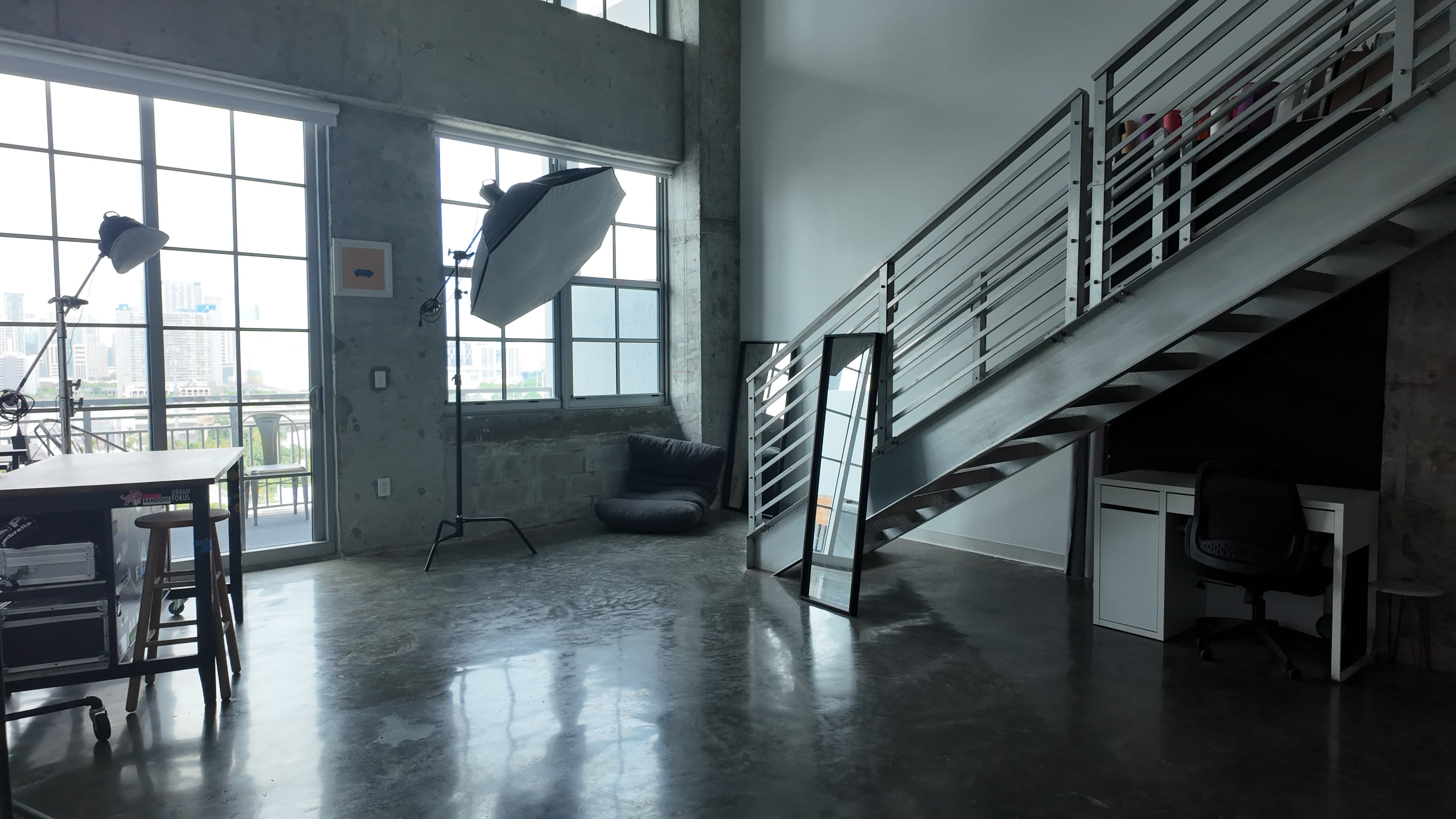 An industrial-style room with large windows, a staircase, a mirror, a desk, and a bean bag chair.