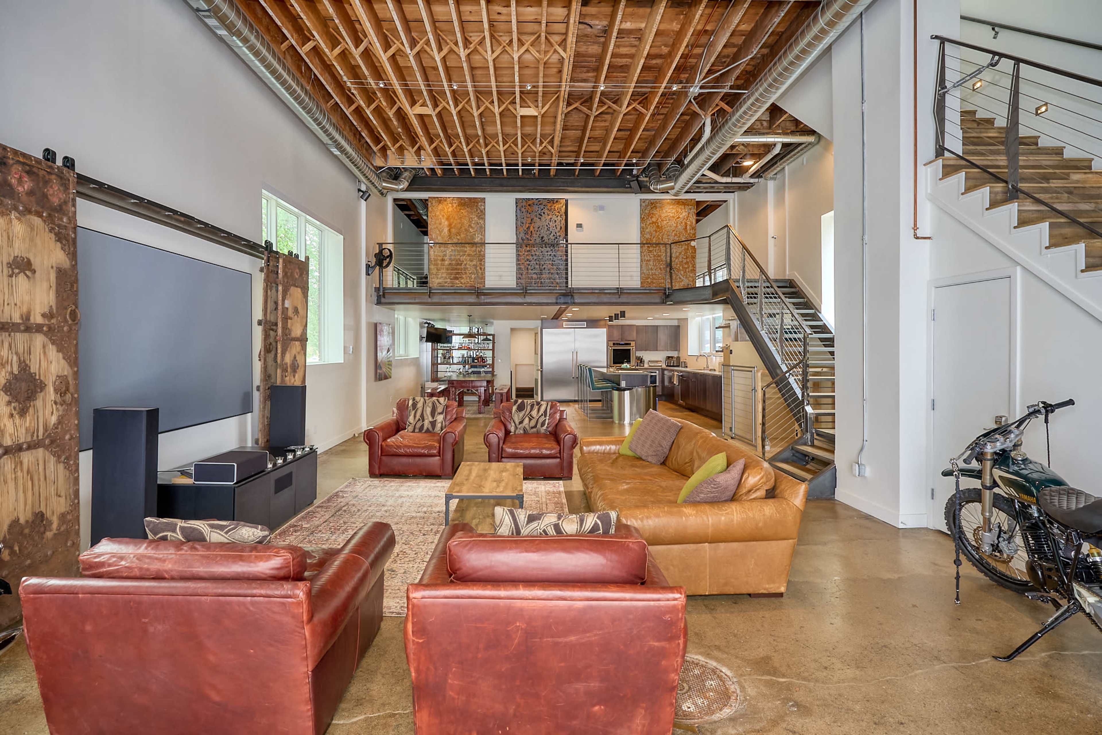 The spacious living area features leather sofas, a large rug, and an open staircase with a kitchen visible in the background.
