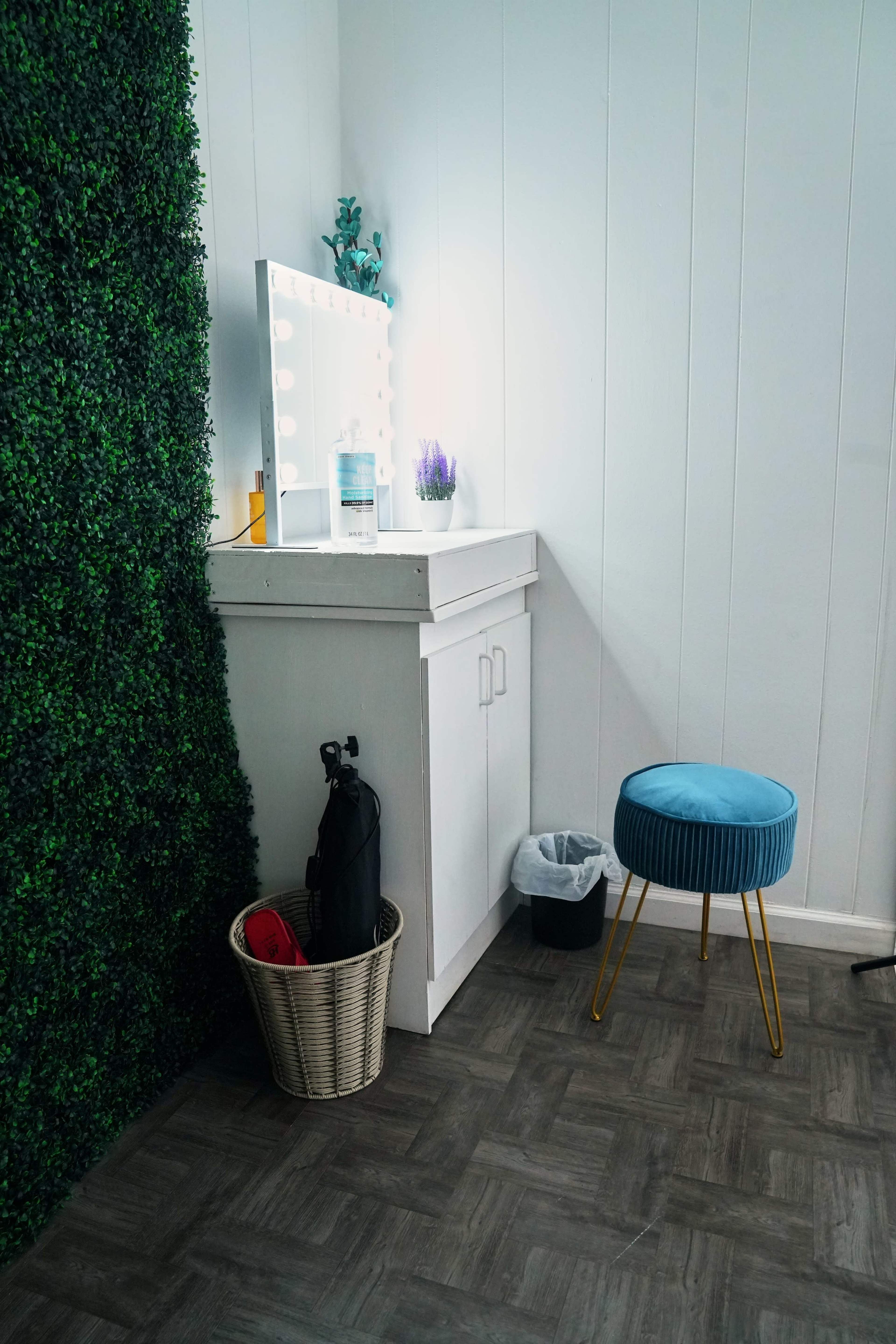 The image shows a small vanity area with a mirror framed by lights, a blue stool, a wastebasket, and a green wall, alongside an umbrella in a basket.