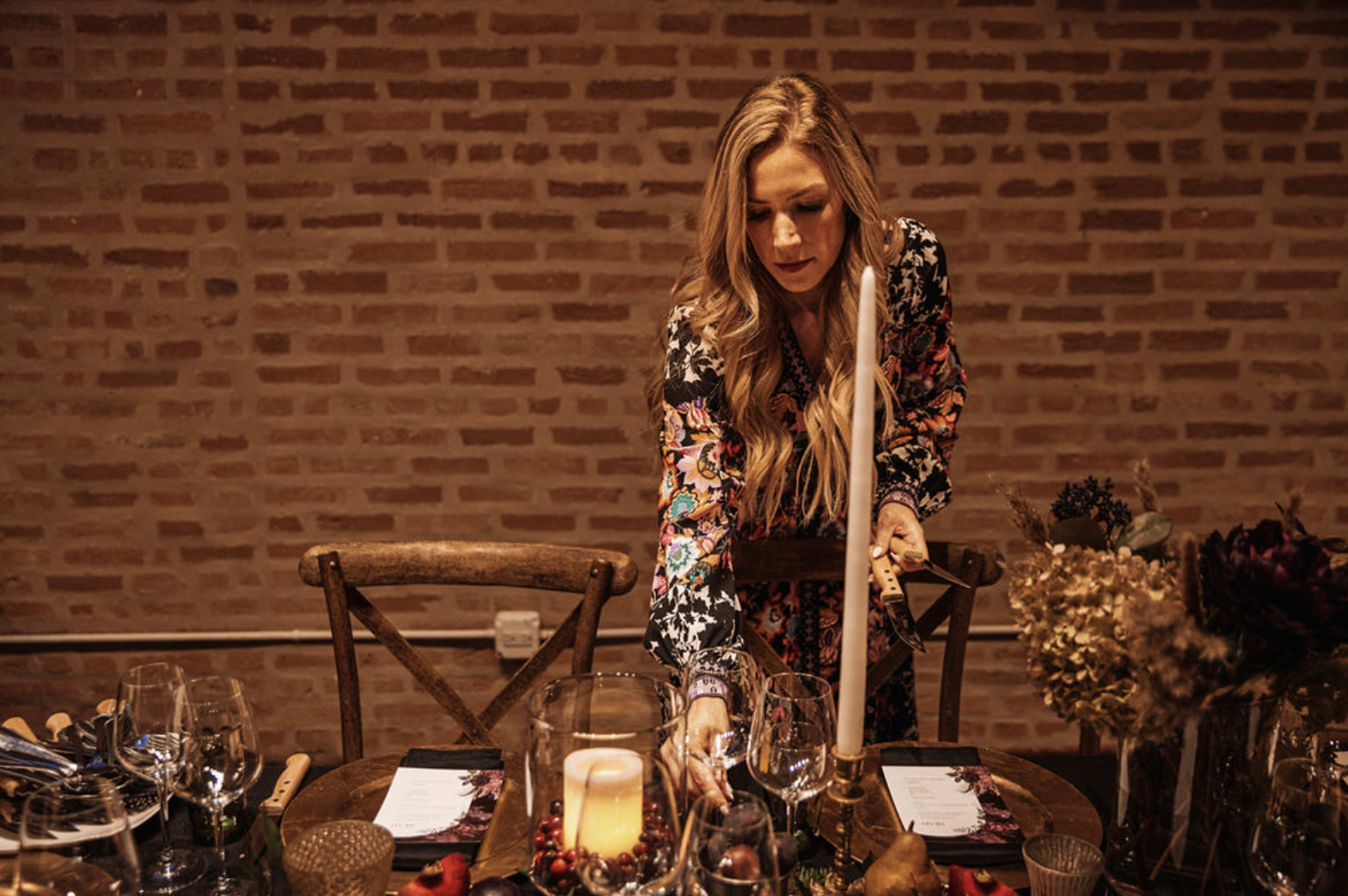 A woman with long hair arranges a table setting with a lit candle and decorative elements in a warmly lit dining space.