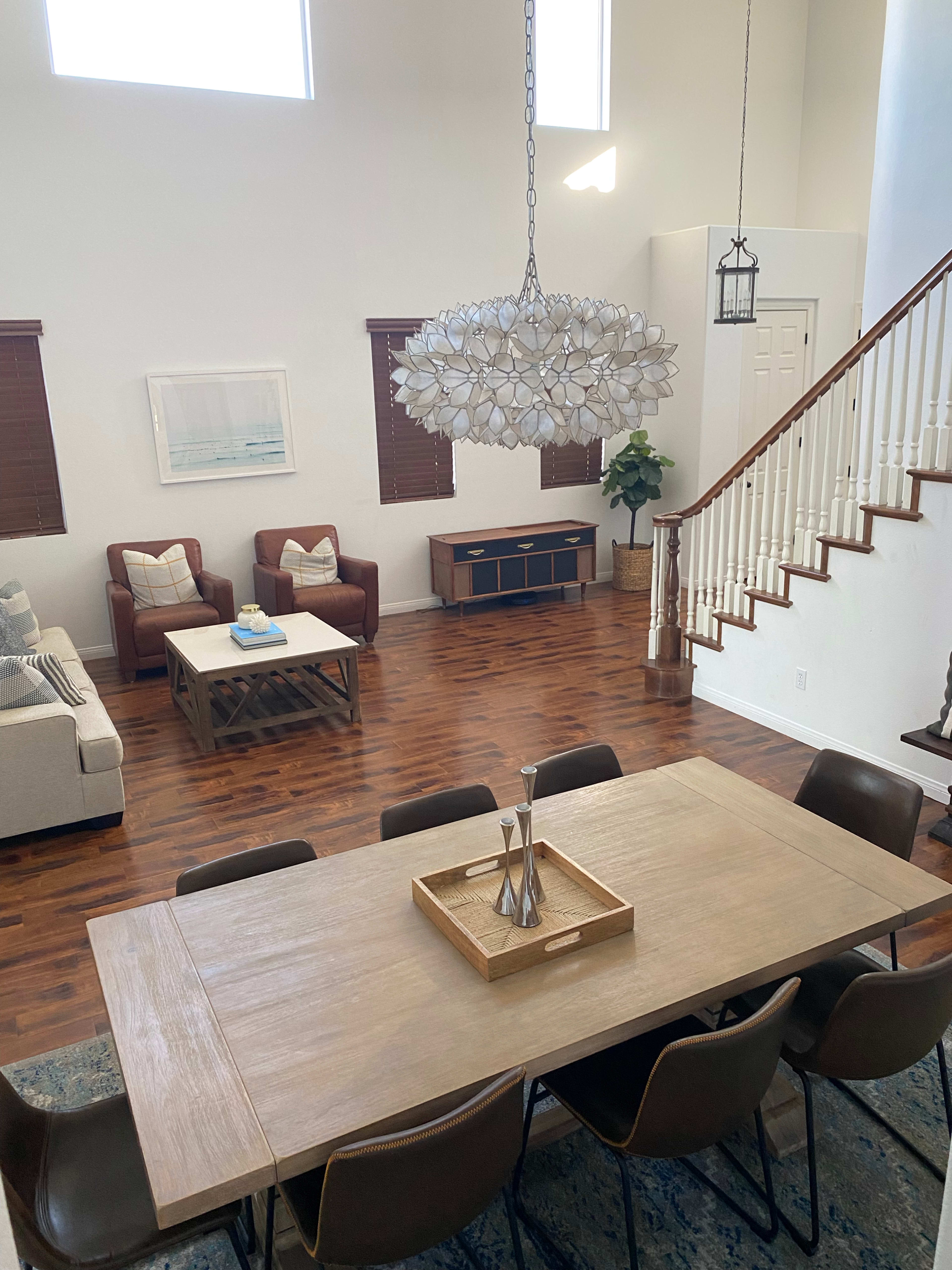A spacious living and dining area features a wooden table, a plush sofa set, and a large chandelier, with sunlight streaming in through the high windows.