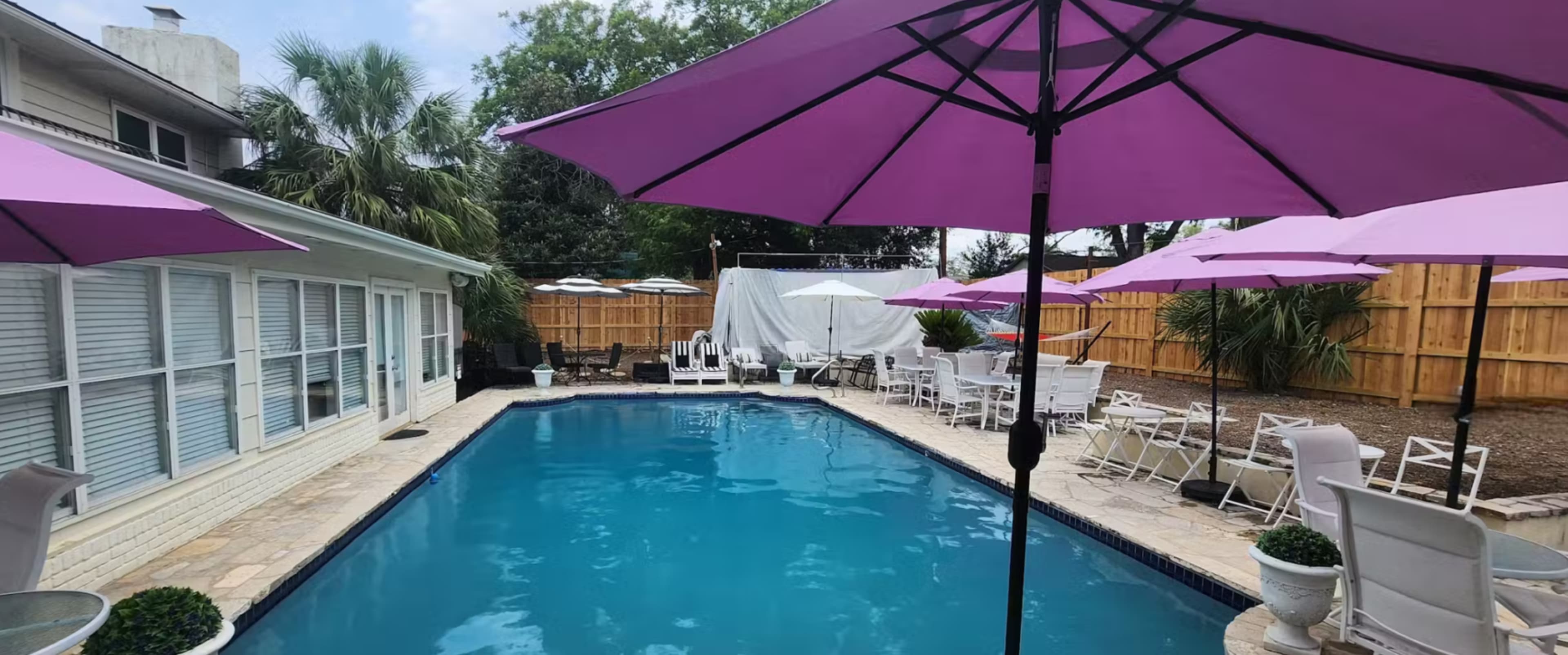 The image shows a swimming pool surrounded by patio chairs and purple umbrellas in a landscaped backyard.