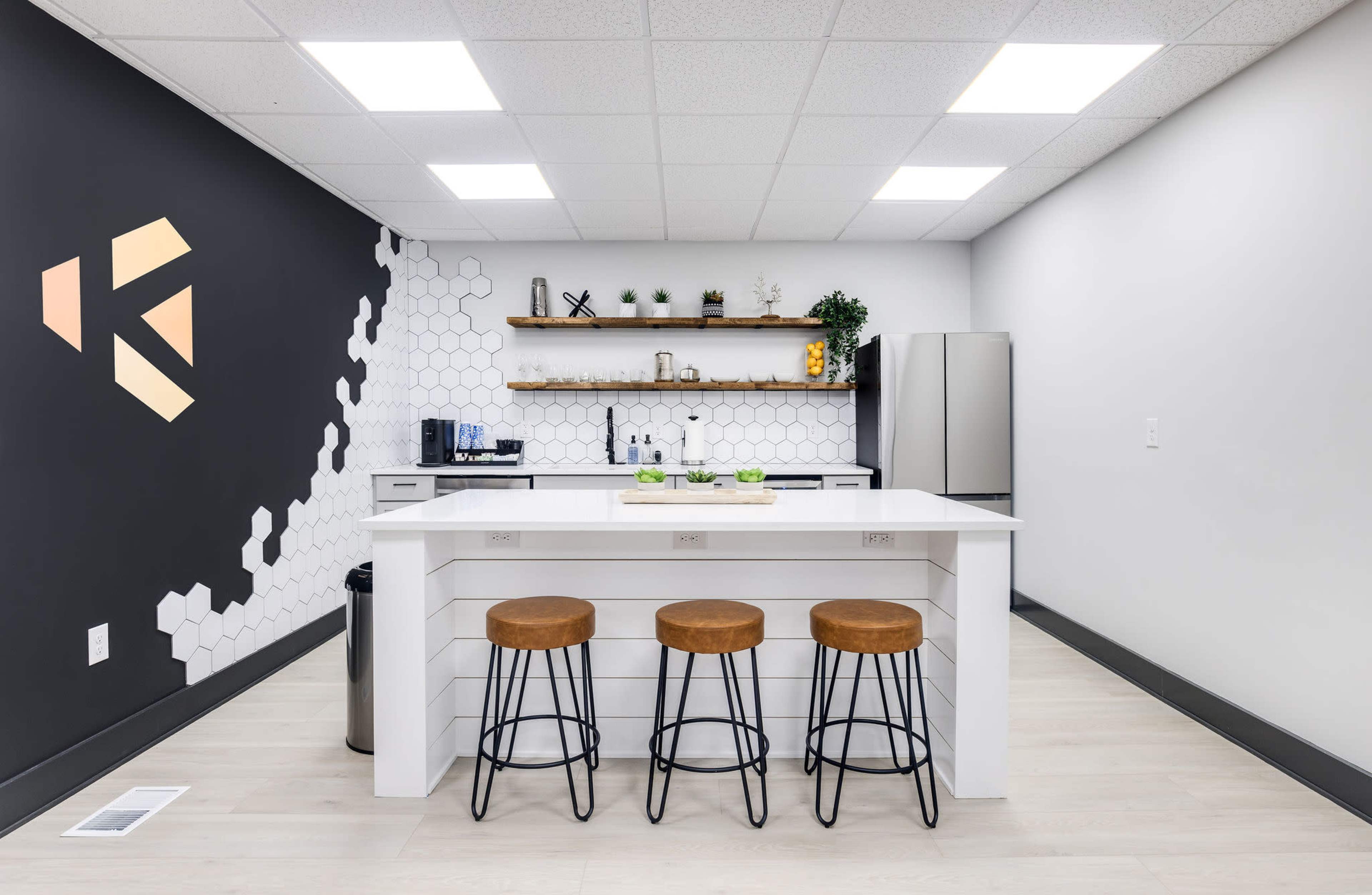 A modern kitchen with a white island, three metal stools, a hexagonal patterned wall, and a sleek silver refrigerator.