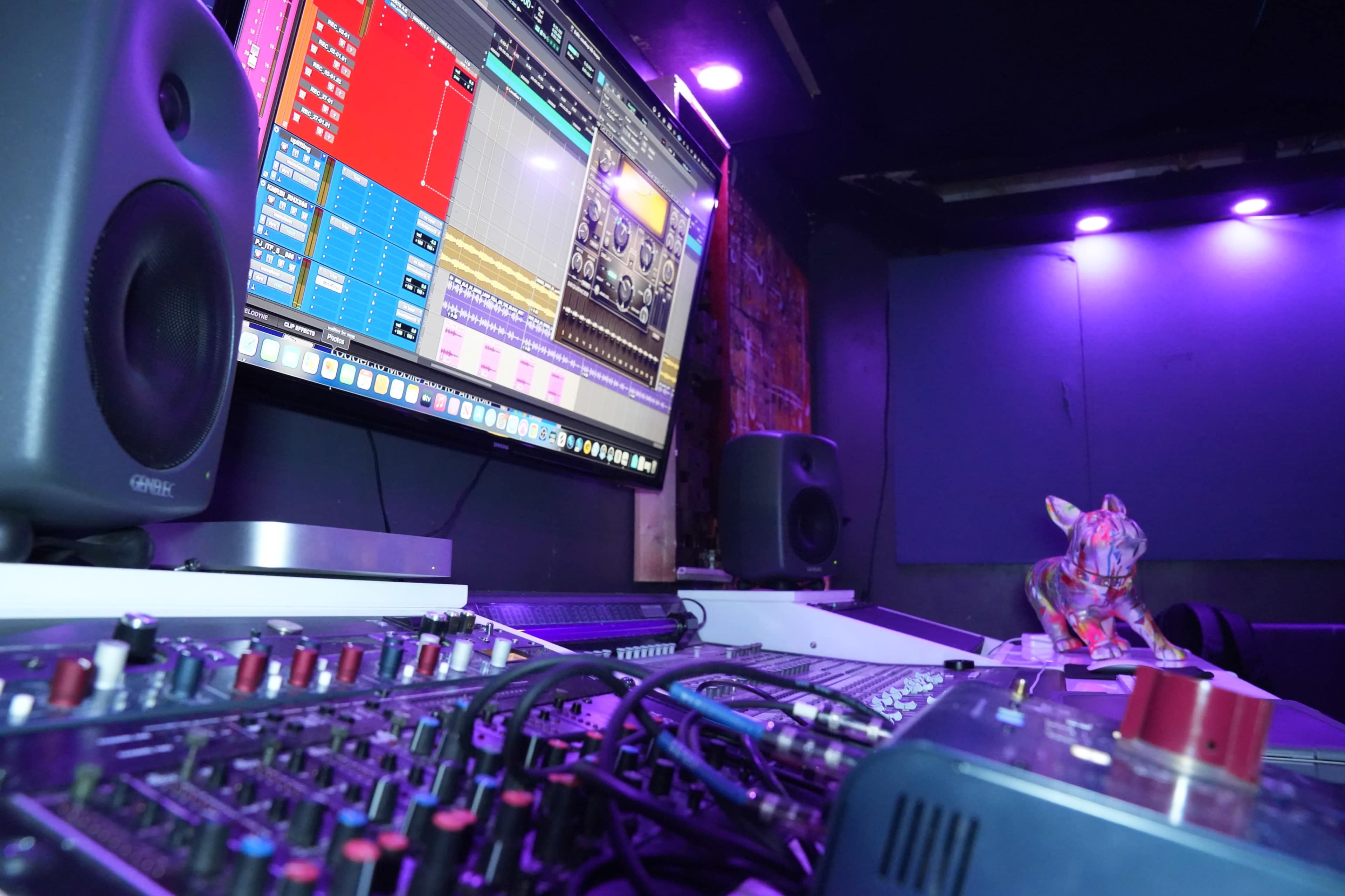 A music production workspace with a large monitor displaying software, audio equipment, and a colorful decorative figurine of a dog.