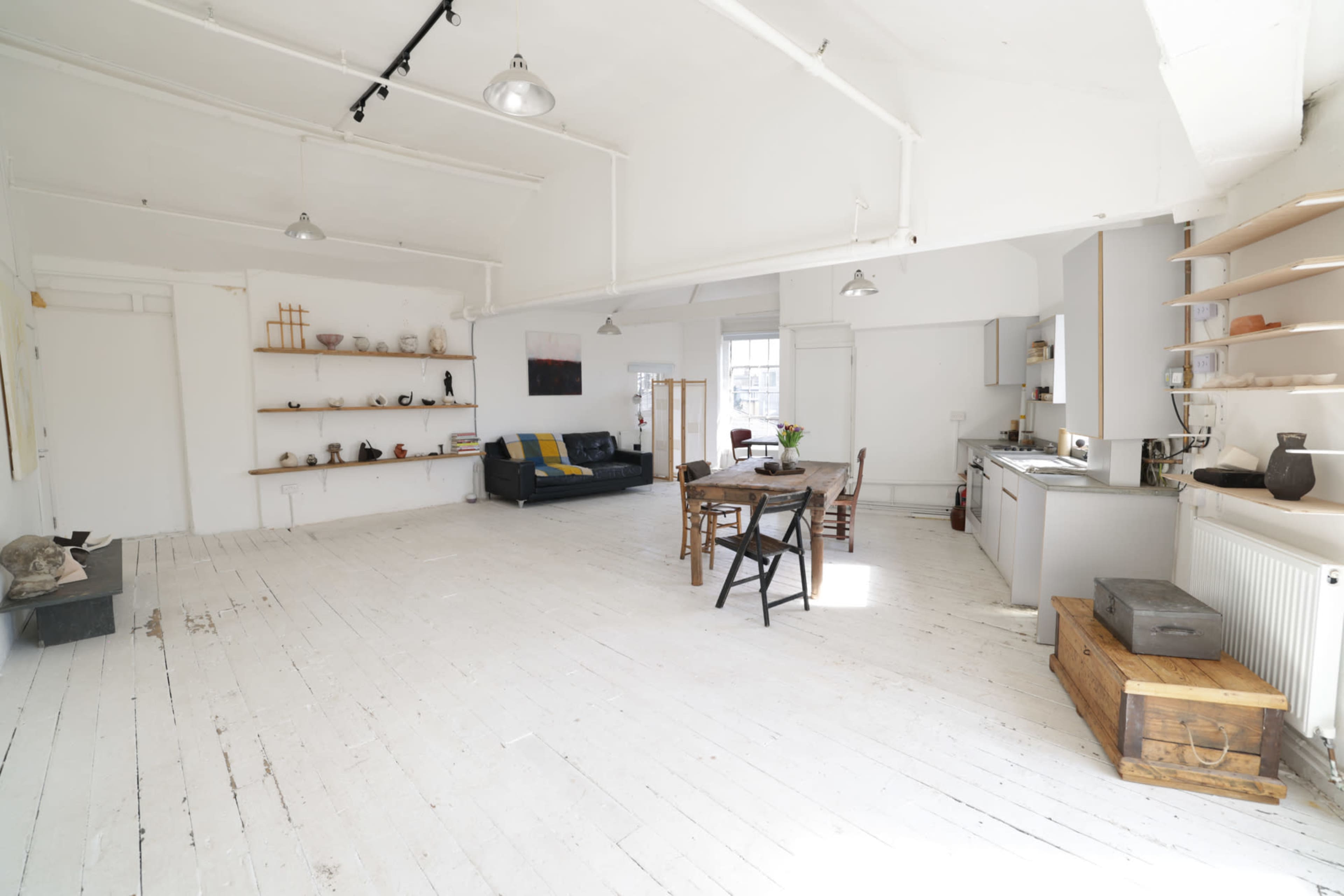 The image shows a spacious, white-painted room with a dining table and chairs, a kitchen area, and shelves displaying various items.