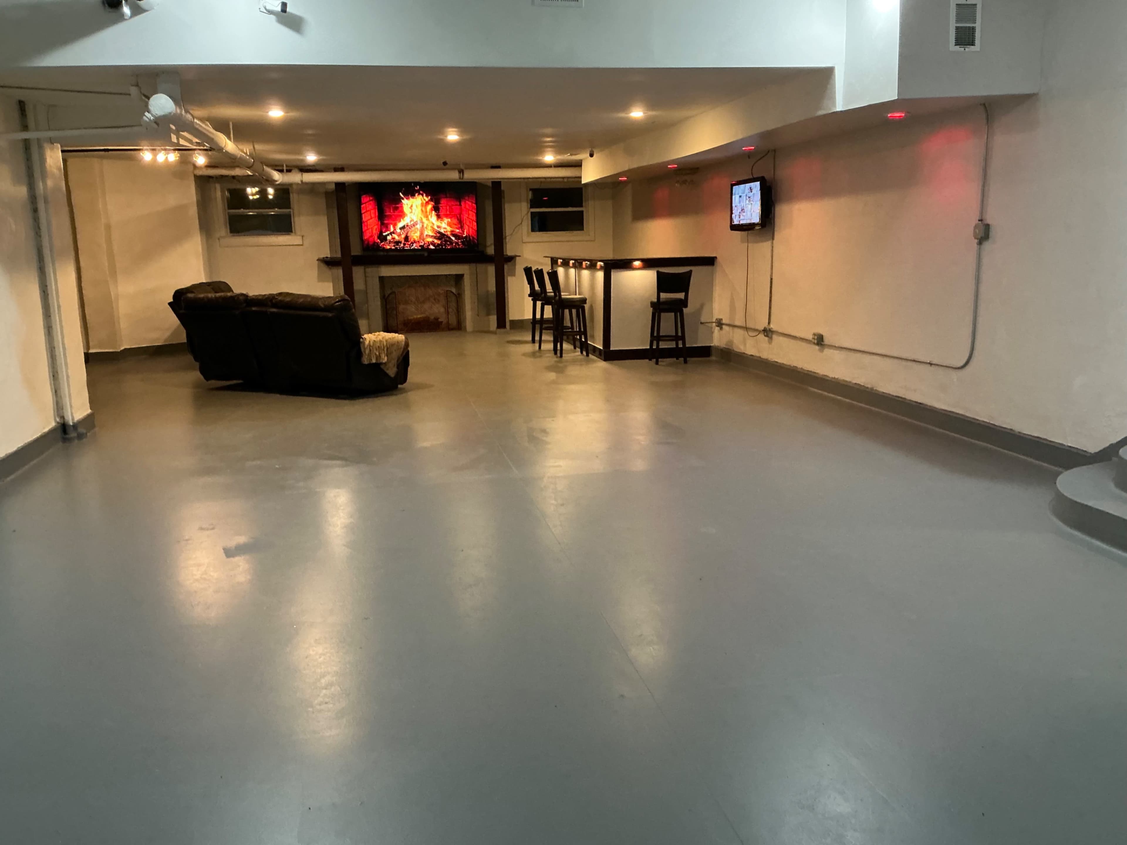 The image shows a spacious, well-lit basement with a seating area, a bar, and two televisions mounted on the walls.