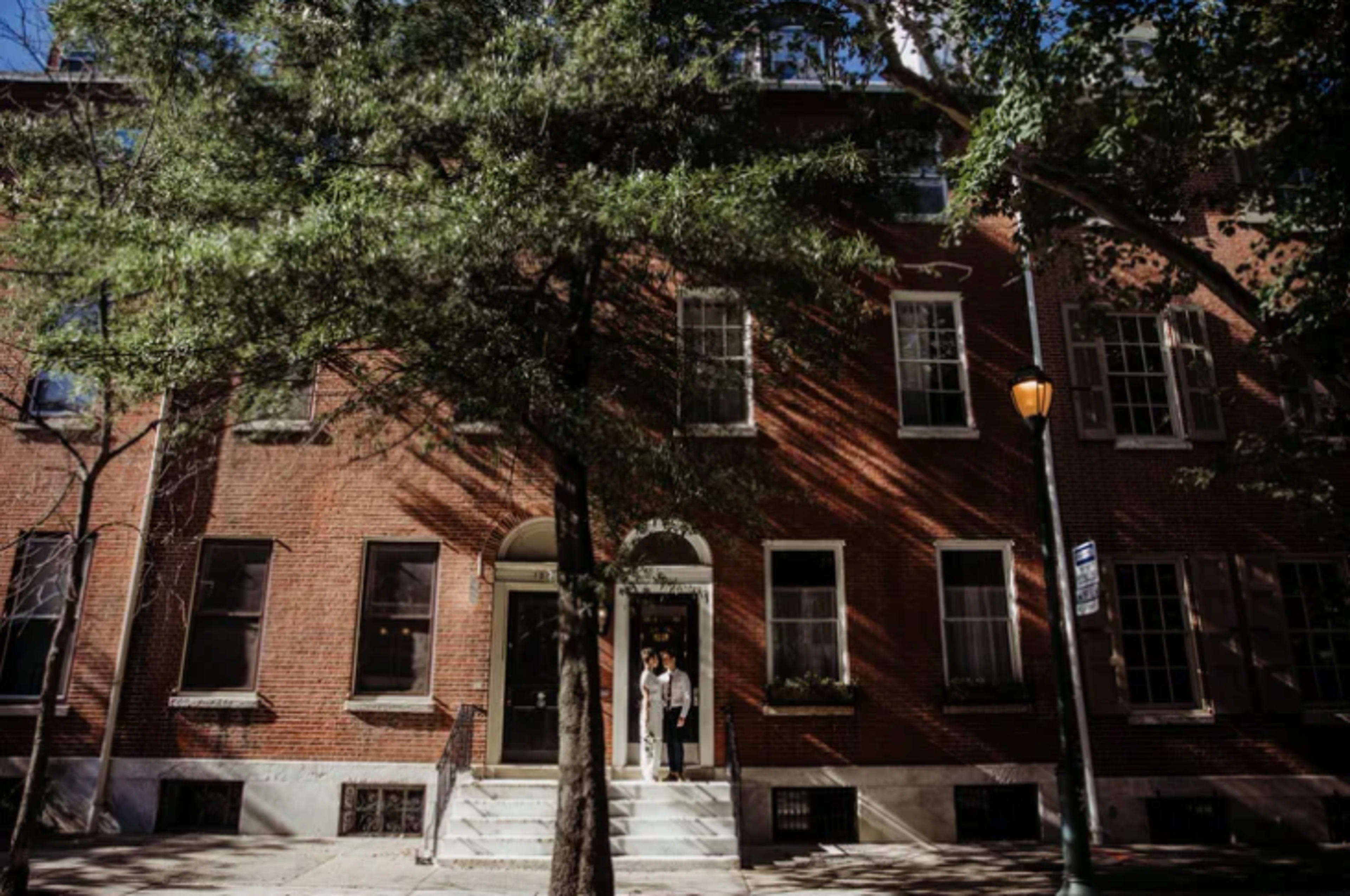 A red brick building with large windows and a set of steps leading to a front entrance, partially shaded by trees.
