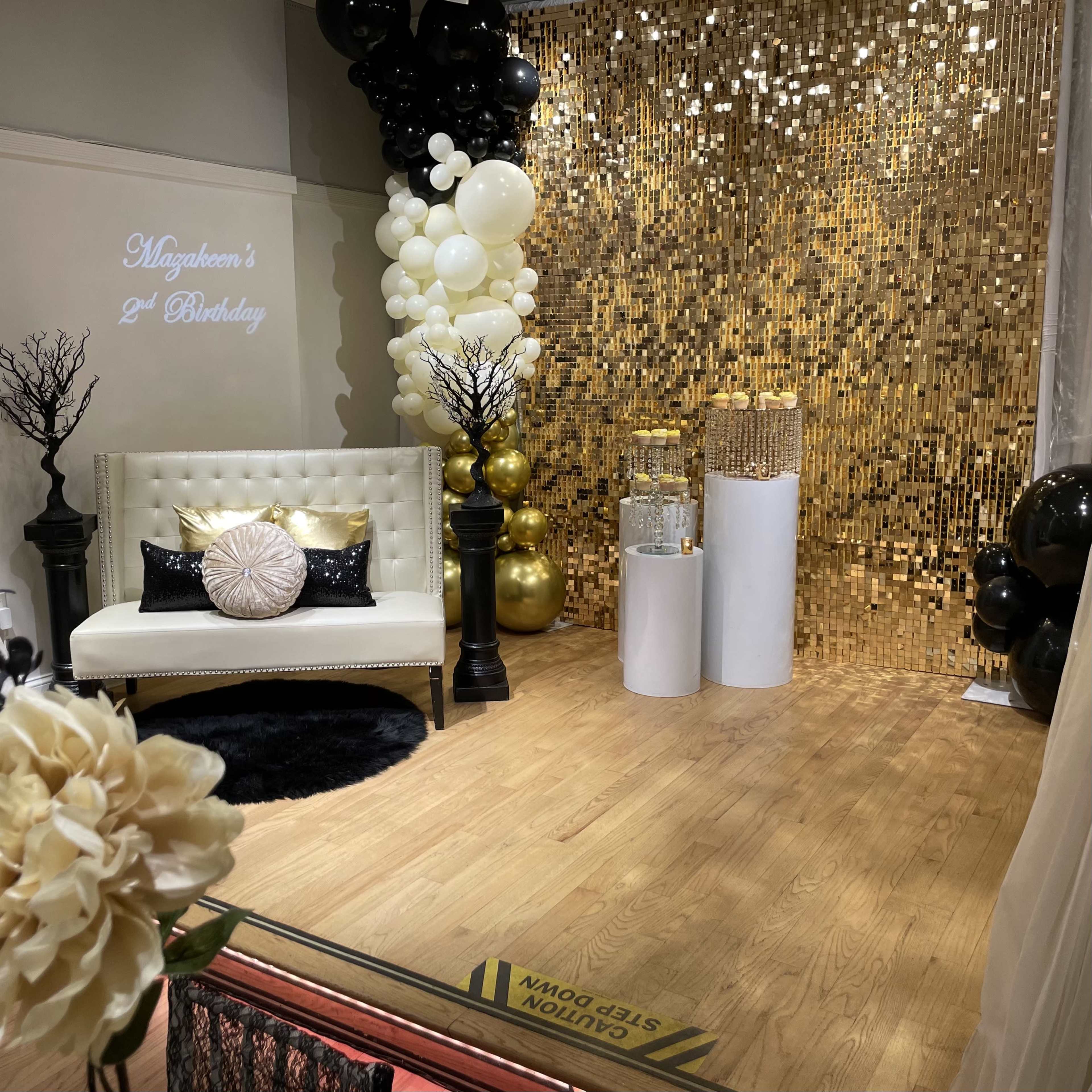 The scene features a decorated event space for a second birthday celebration, with a gold backdrop, a white sofa, and black and gold balloons.