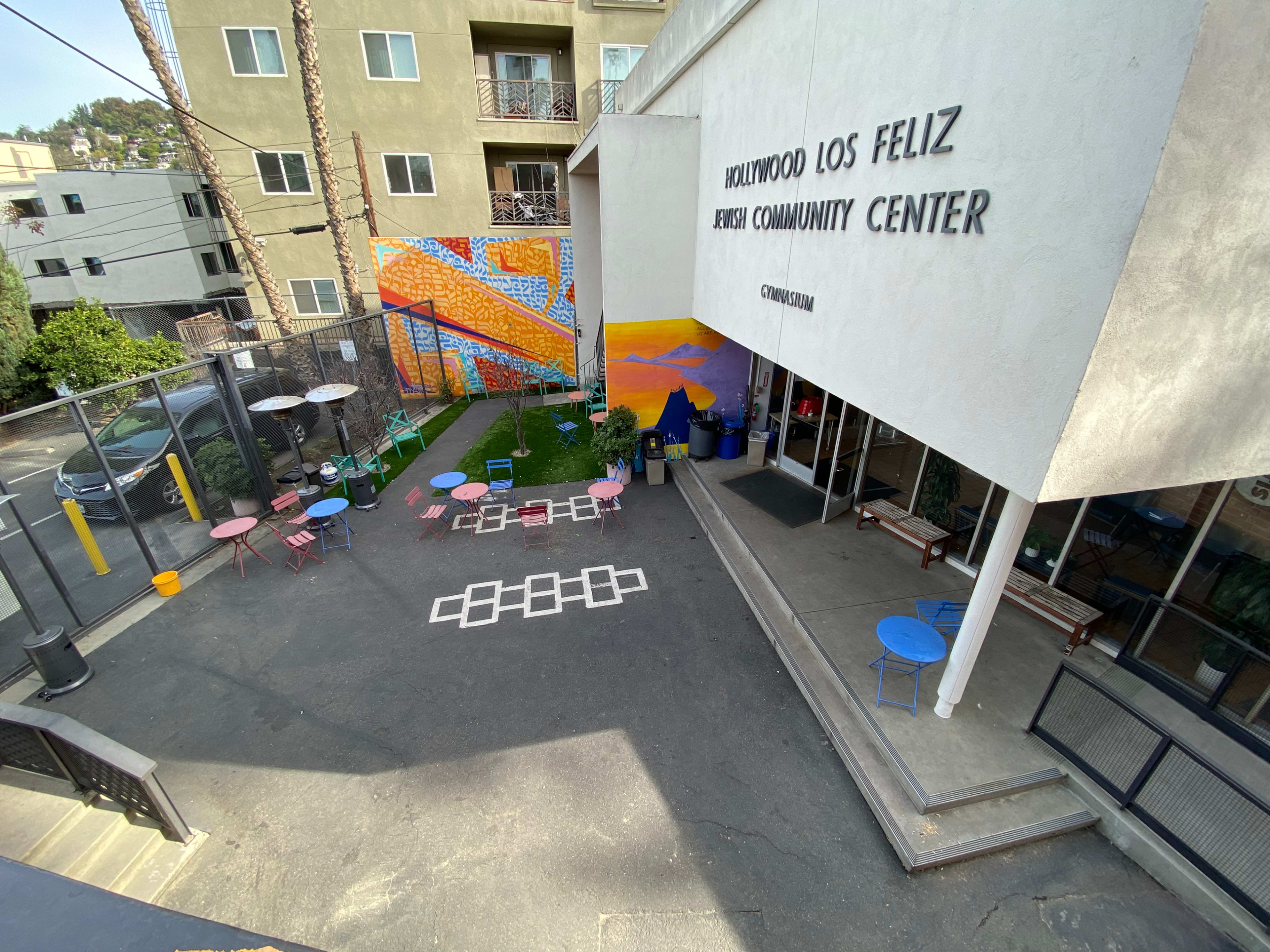 The image shows the exterior of the Hollywood Los Feliz Jewish Community Center, featuring a colorful mural and outdoor seating arrangements in a courtyard.
