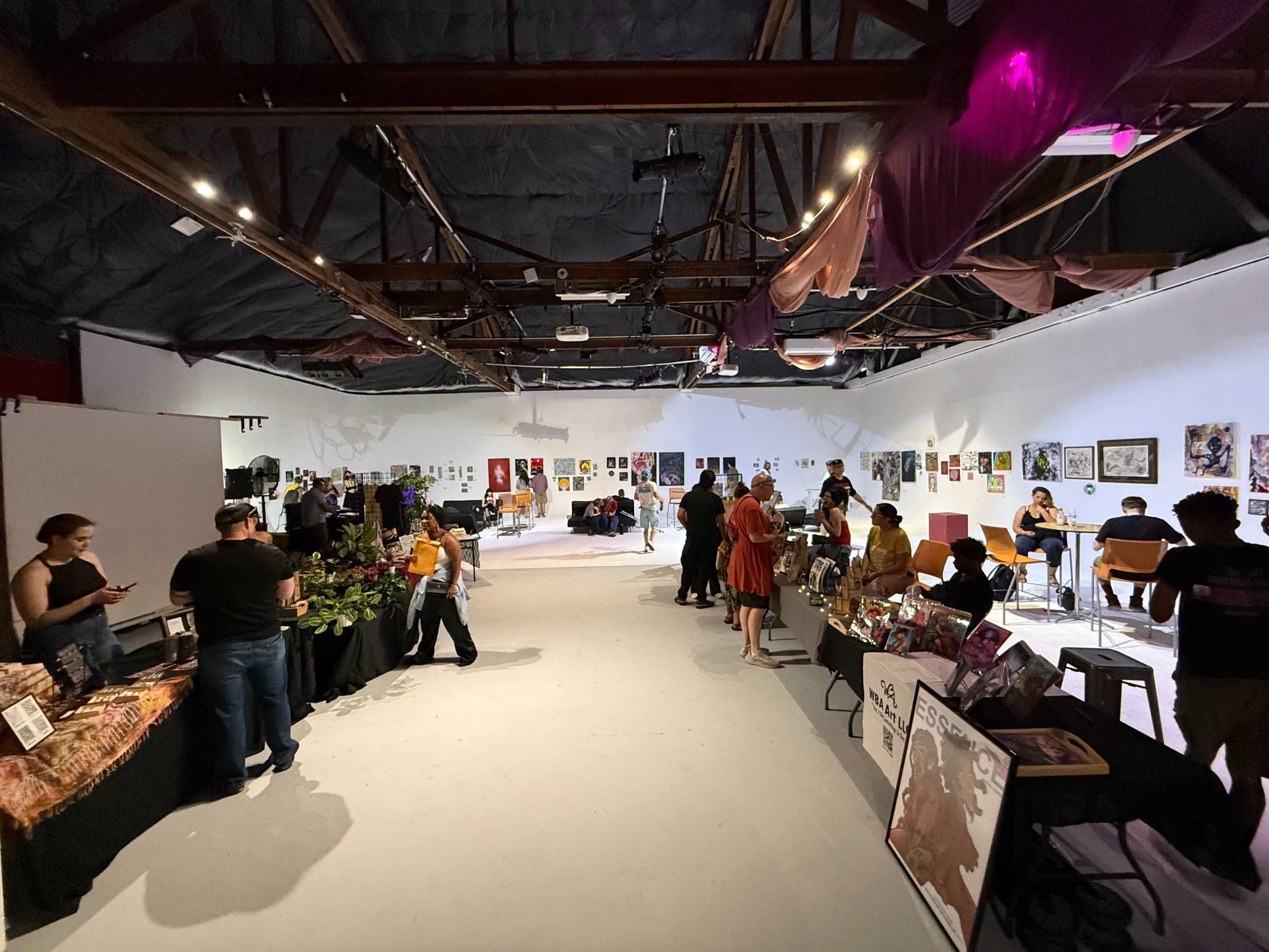 The image shows an art exhibition space with various tables displaying artwork, vendors selling items, and people engaging in conversation throughout the room.