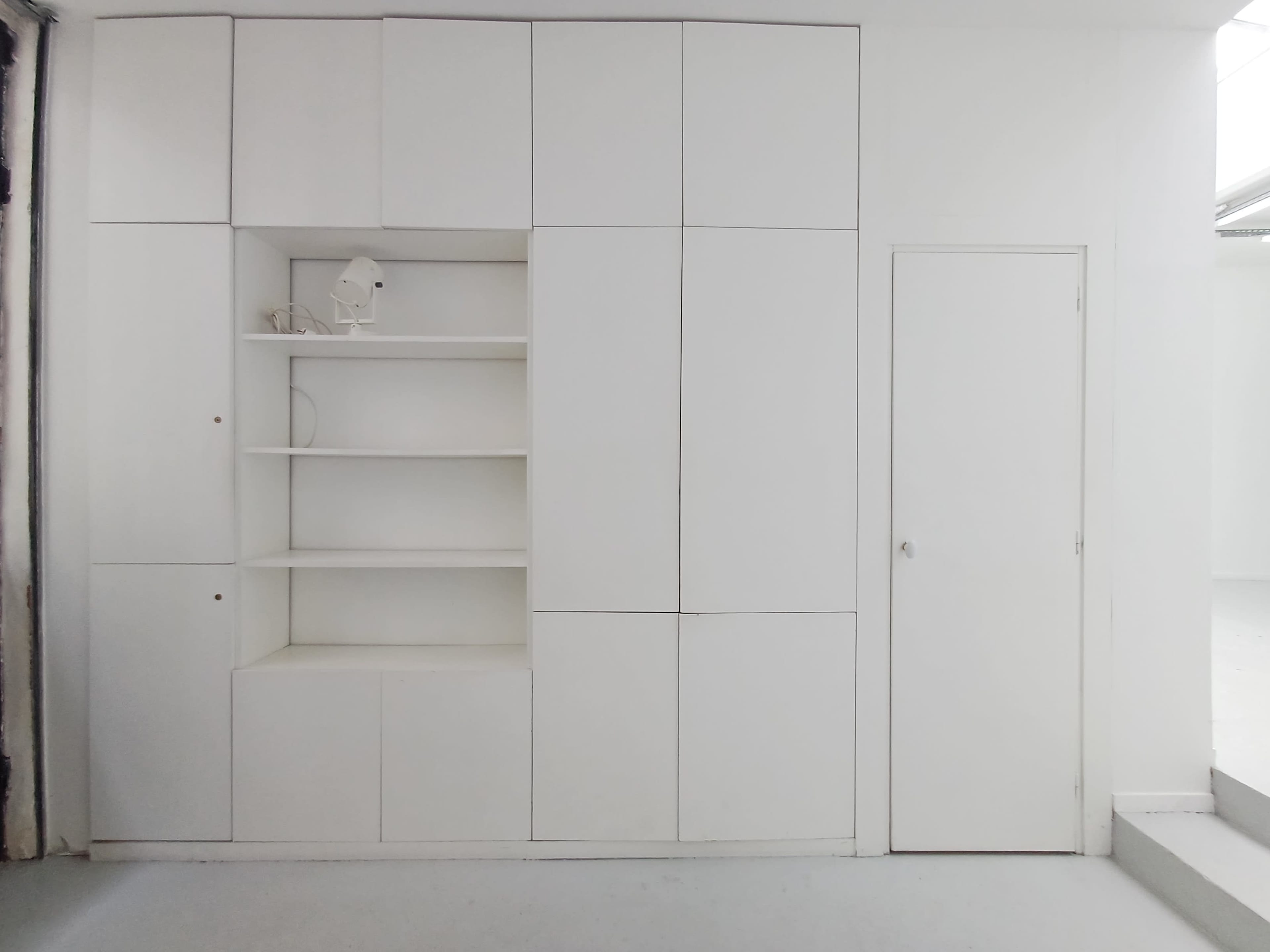 The image shows a white wall unit with shelves and a door, set against a plain white background.