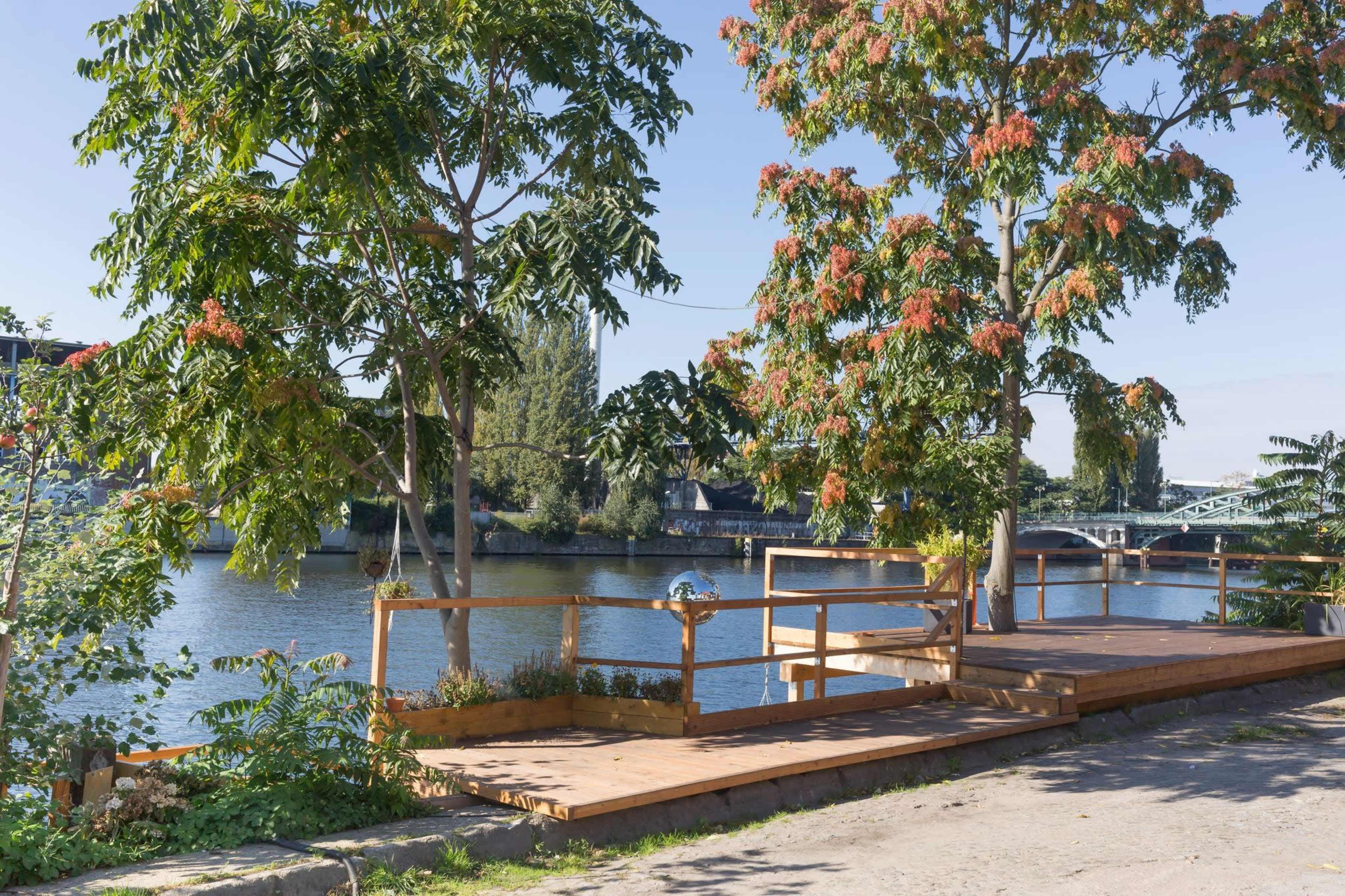 The image shows a riverside walkway with wooden decking and trees, alongside a calm body of water and a bridge in the background.