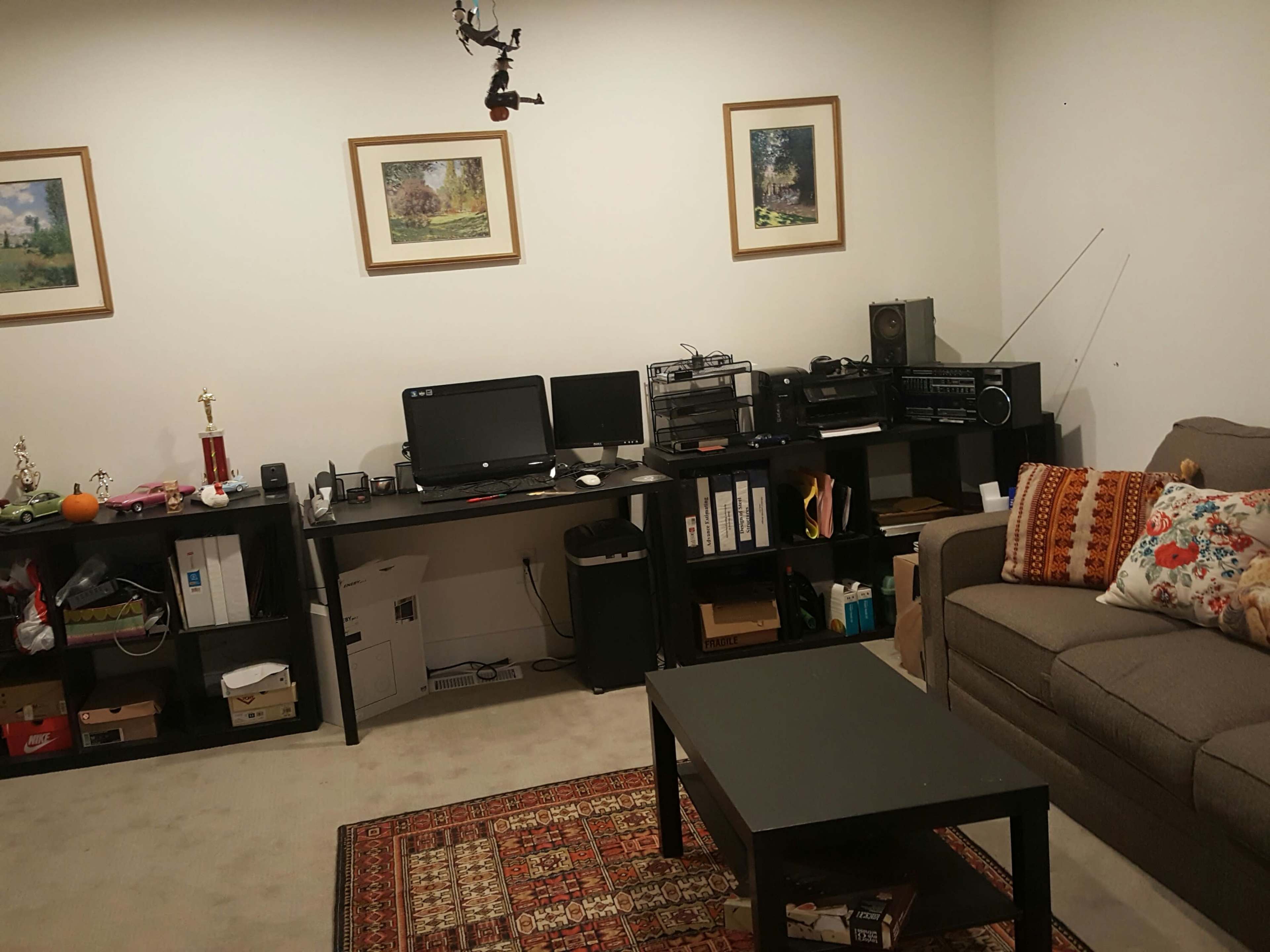 A living room features a sofa, a coffee table, a desk with two computer monitors, and shelves containing various items, including a trophy and framed pictures on the walls.