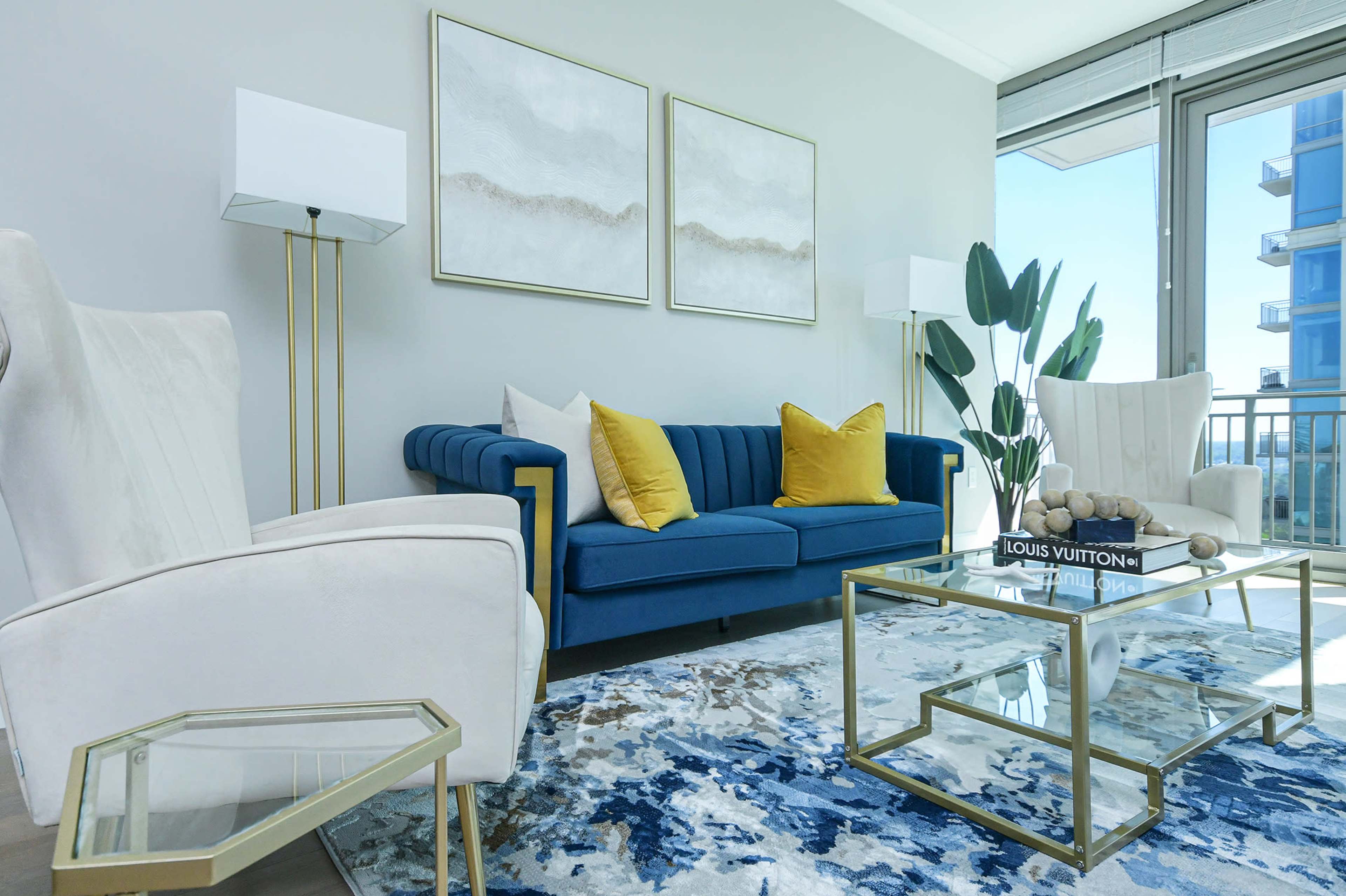 A modern living room with a blue sofa, two white armchairs, a glass coffee table, and decorative plants, all set against a light gray wall.