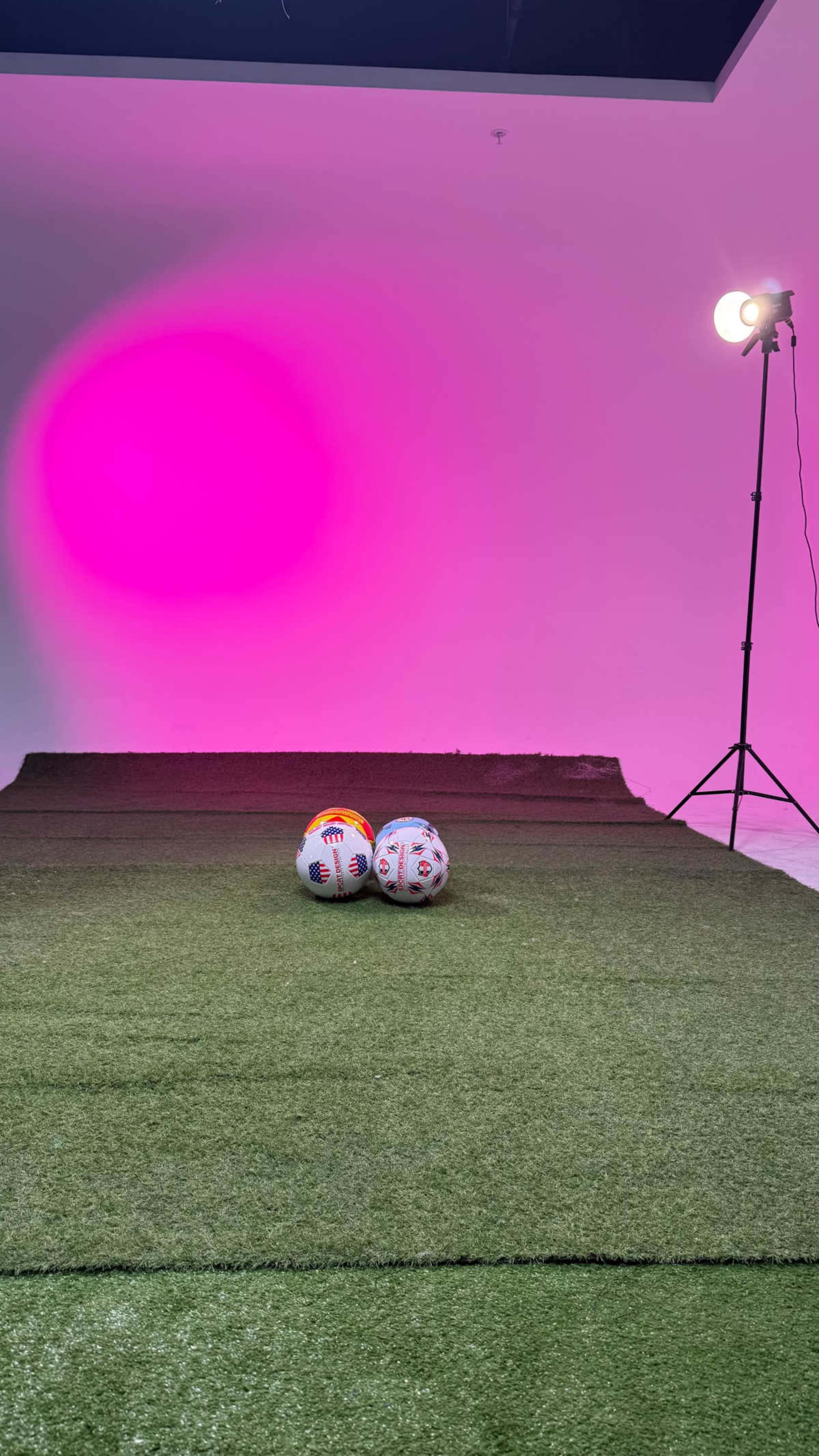 Two soccer balls are placed on a patch of artificial grass under a pink spotlight.