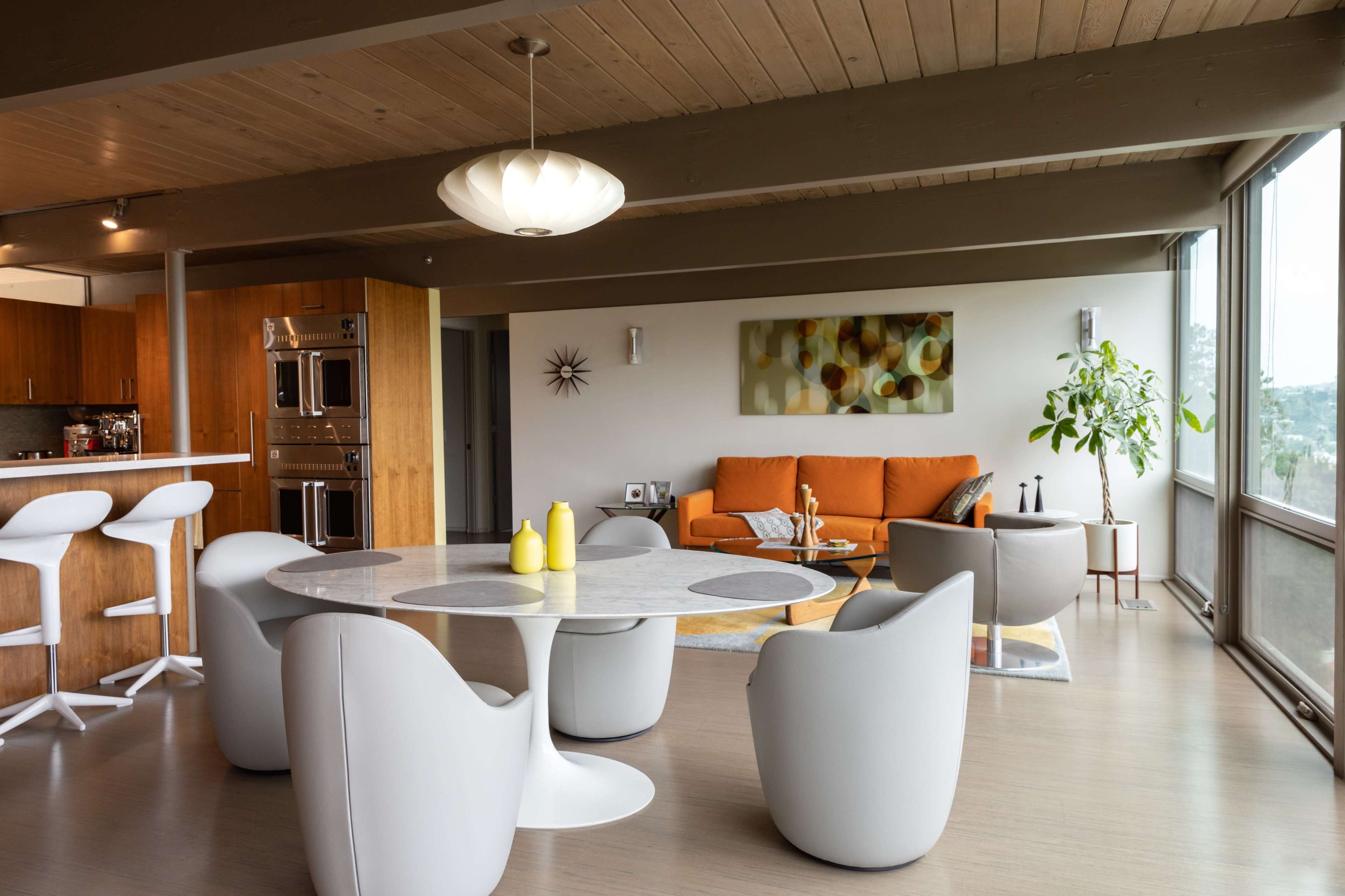A modern living space features a round dining table with white chairs, an orange couch, and large windows allowing natural light to fill the room.