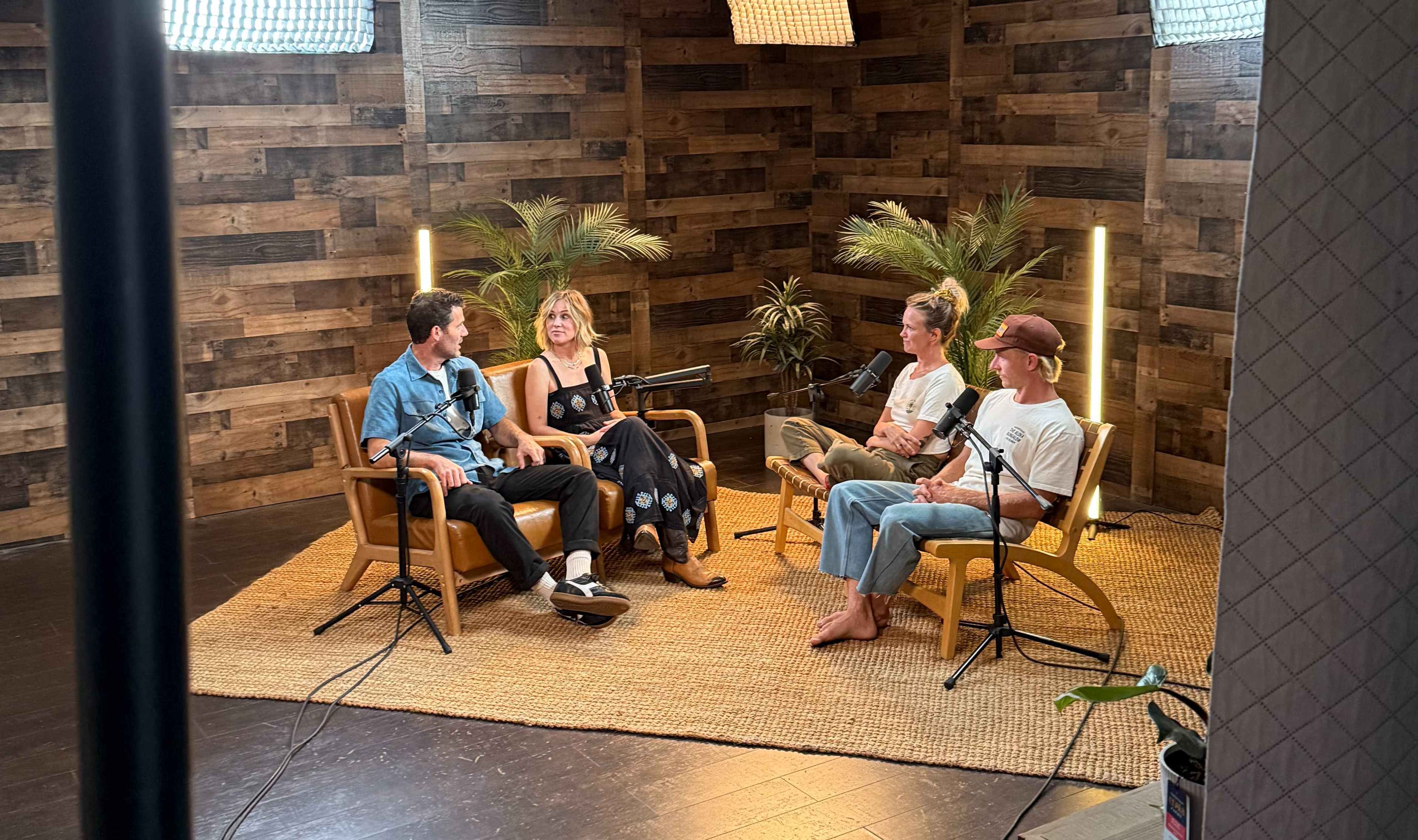 A group of four people sits in a studio setting with wooden walls, engaged in conversation around microphones.