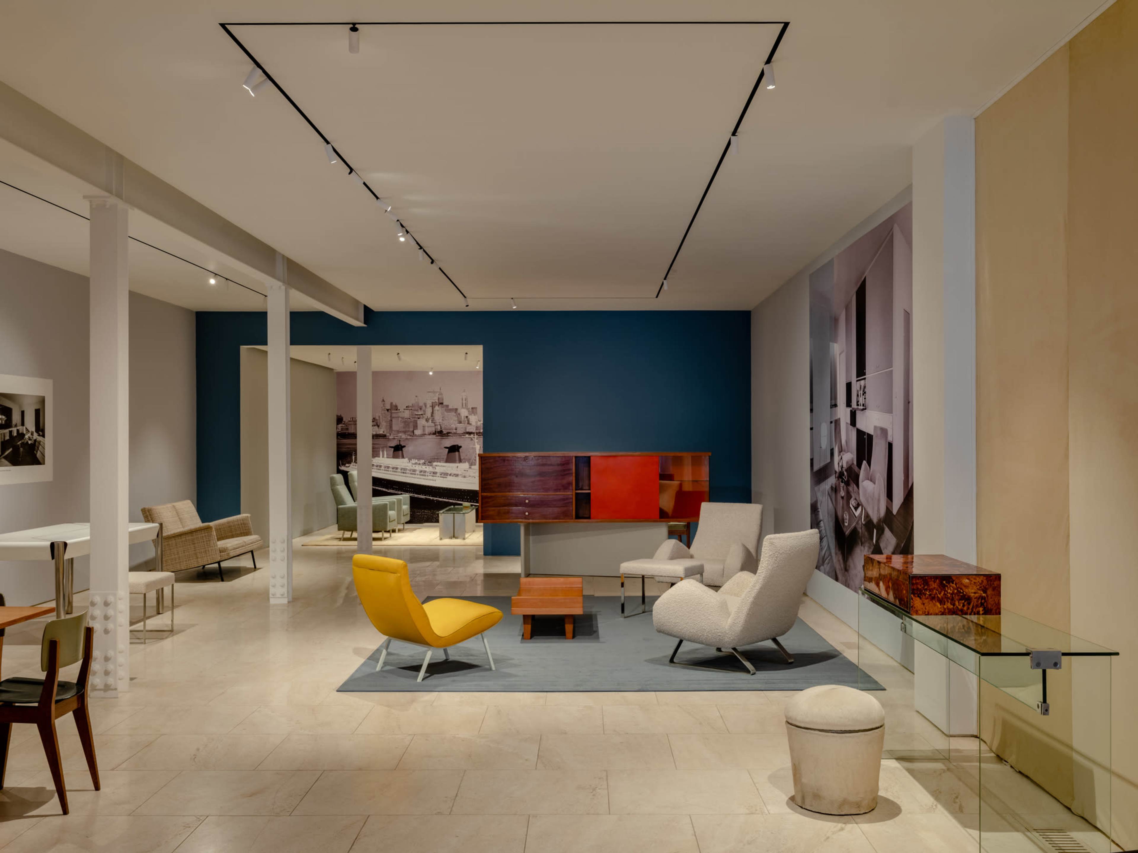 The image shows a modern, minimalistic interior space featuring various furniture pieces, including armchairs, tables, and a display case on a light-colored floor.