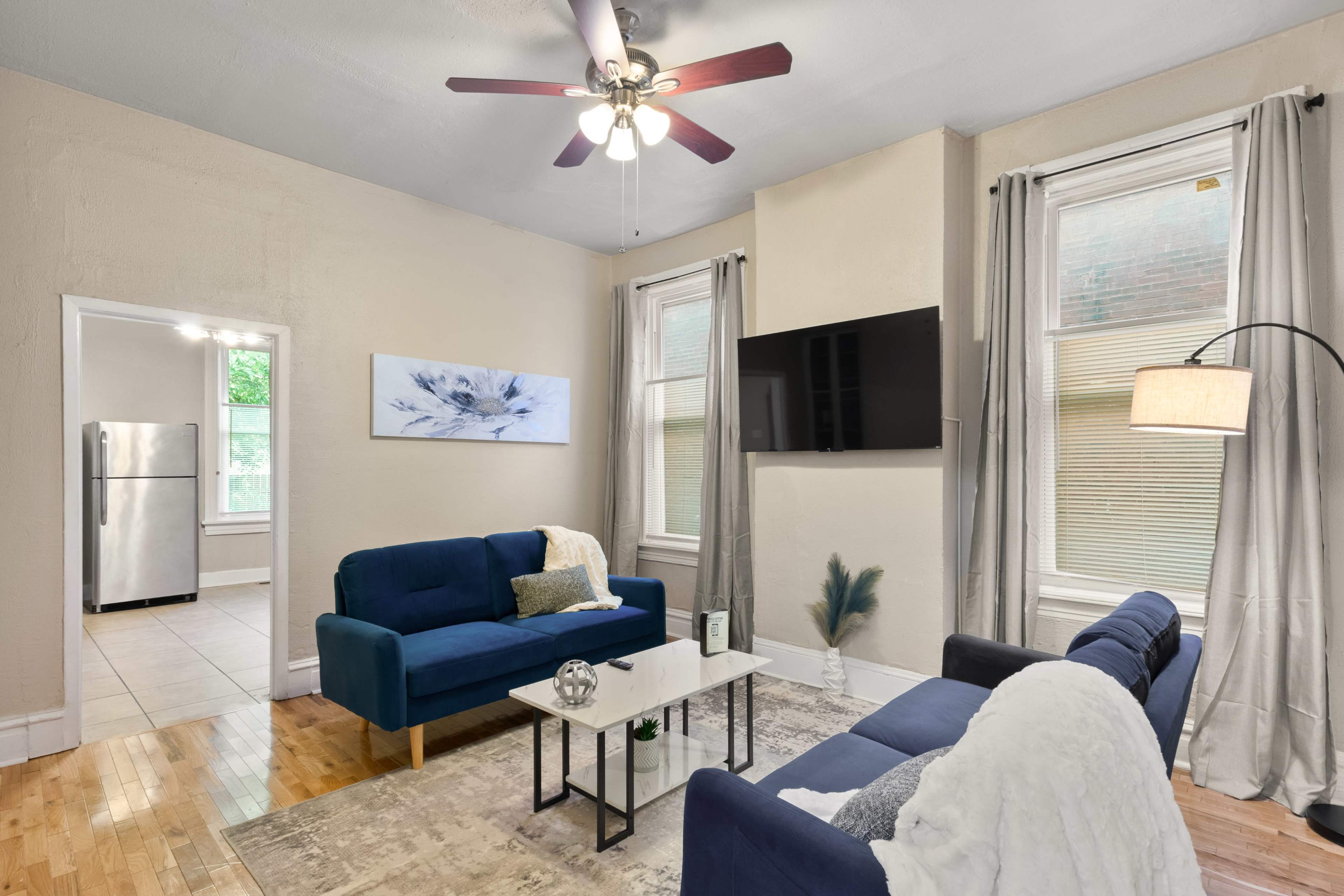 A living room with two blue sofas facing a coffee table, a wall-mounted TV, and a doorway leading to a kitchen in the background.