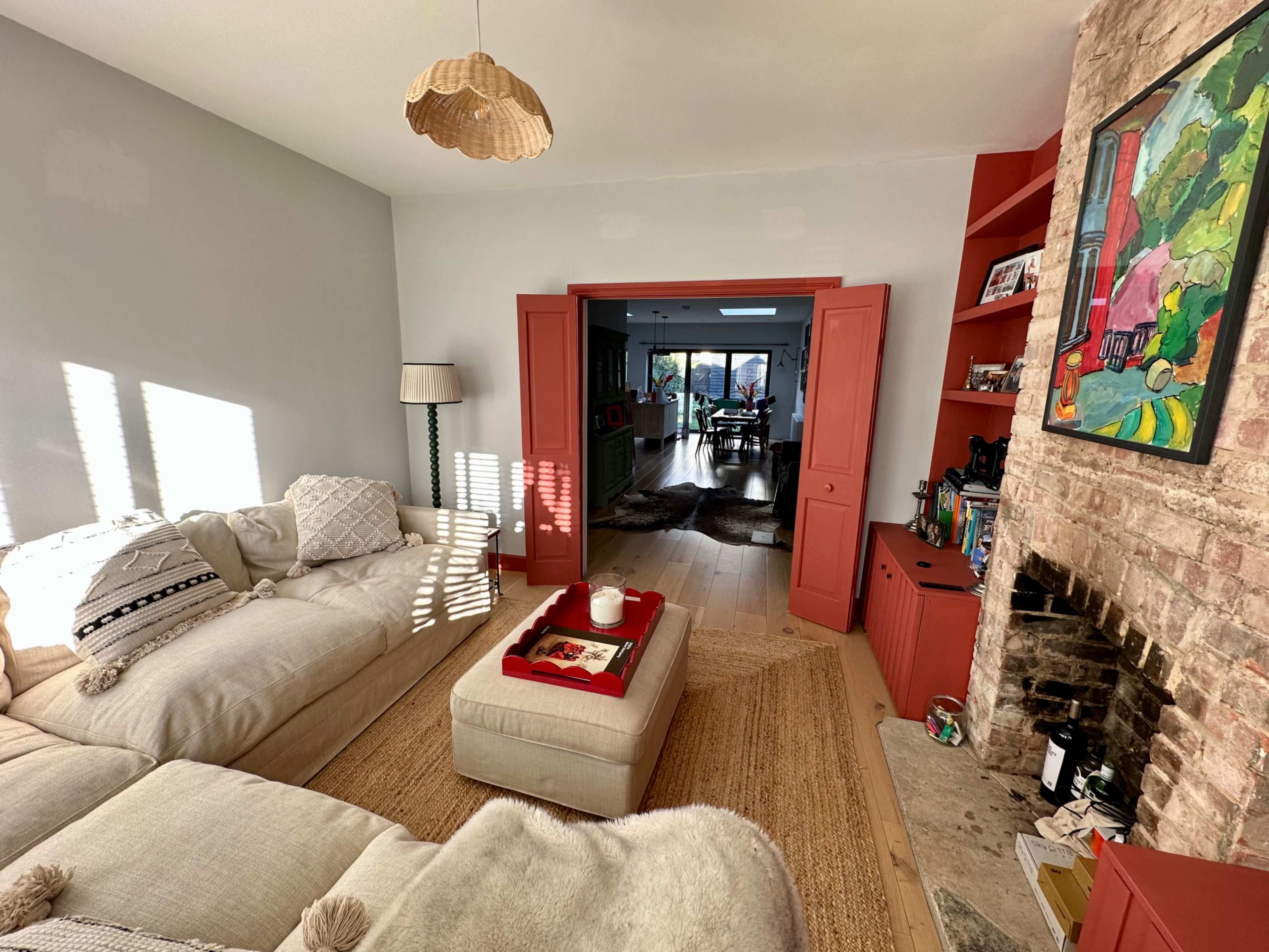 The image shows a modern living room with a large light-colored sofa, a coffee table on a rug, and a cozy fireplace, while an open doorway leads to a brighter area with large windows.