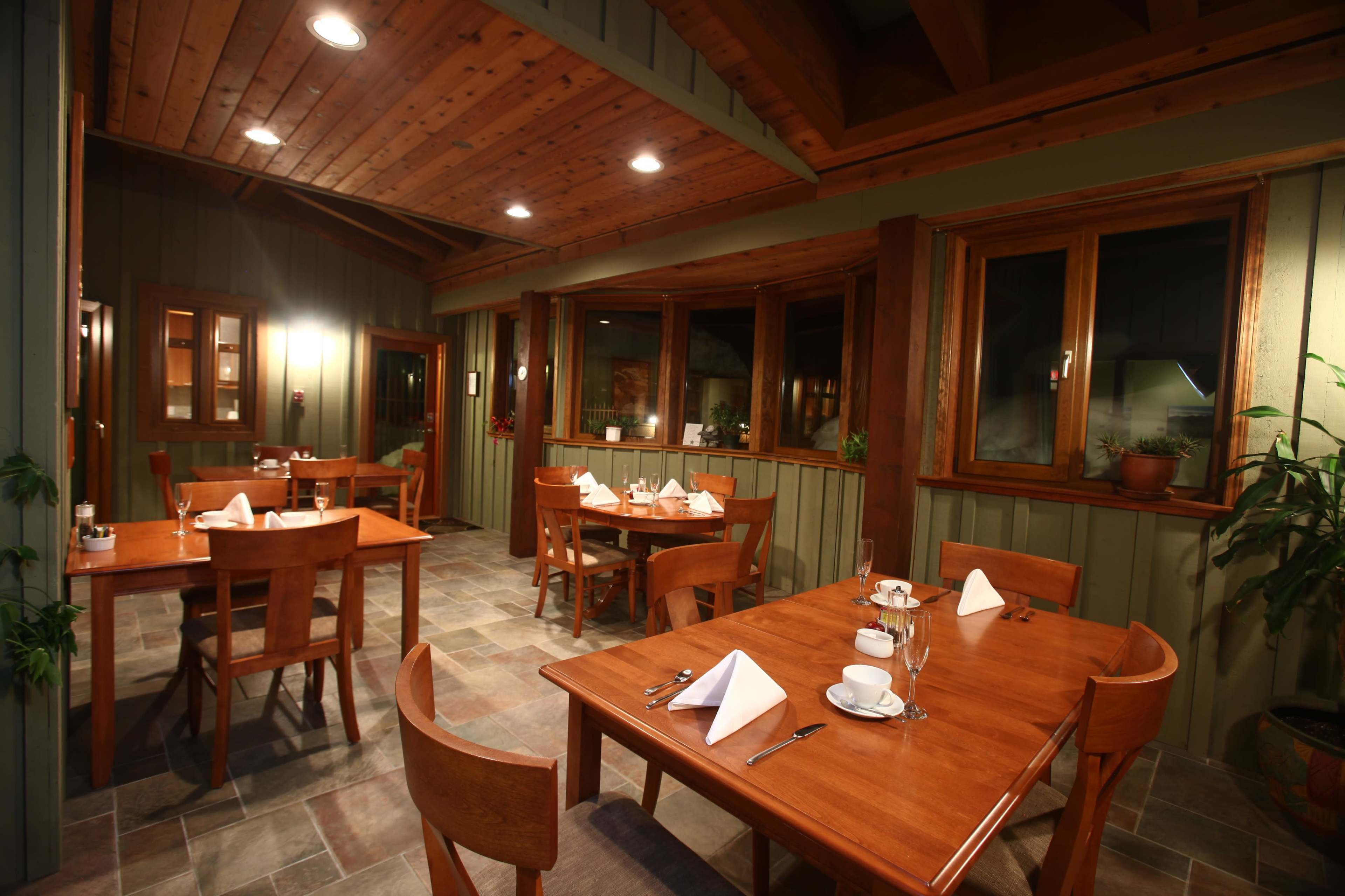 The image shows a rustic dining area with wooden tables set for meals, surrounded by green and brown walls, and large windows letting in light.