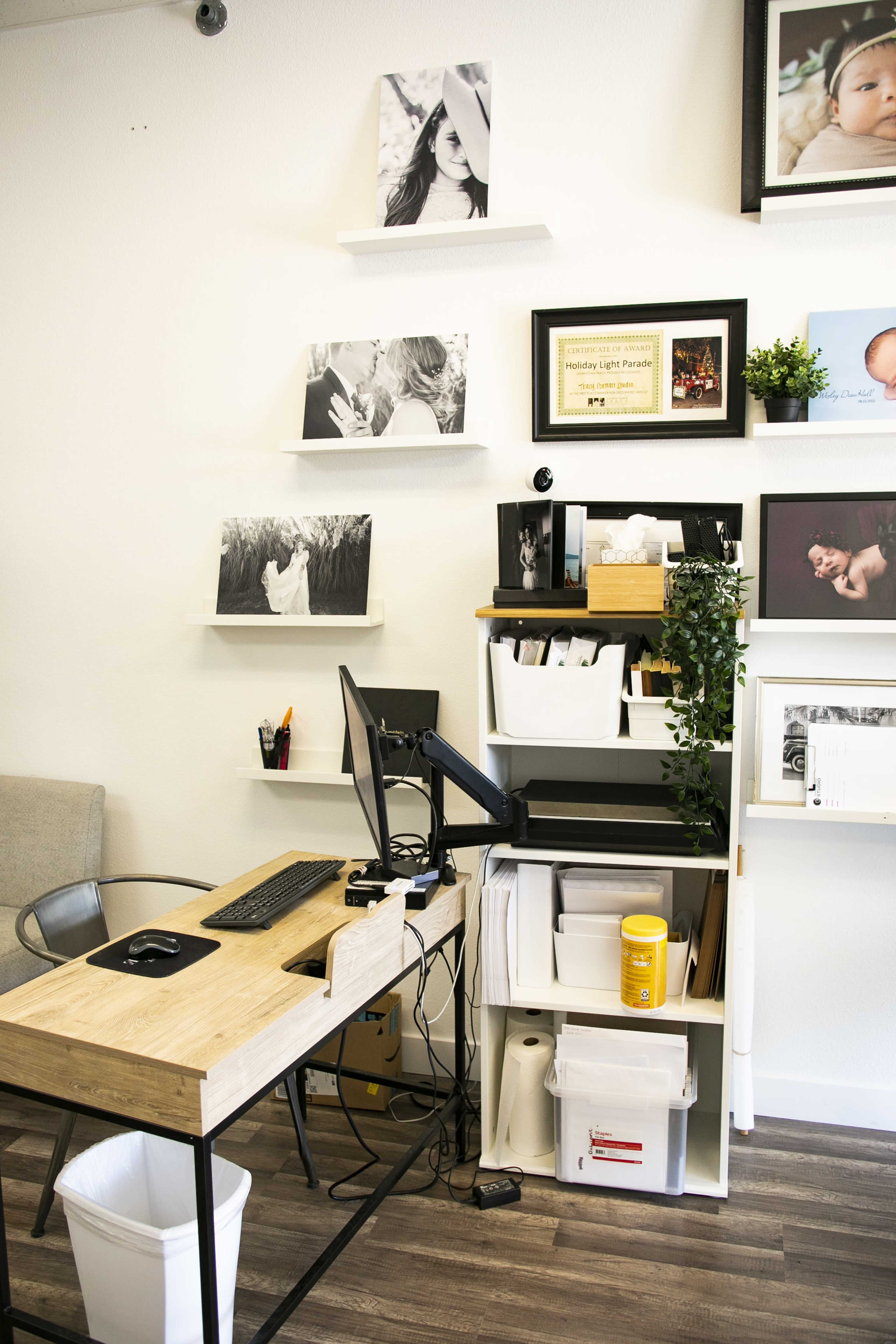 The image shows a small office workspace featuring a wooden desk with a computer, shelves displaying photographs and a certificate, and storage units with various supplies.