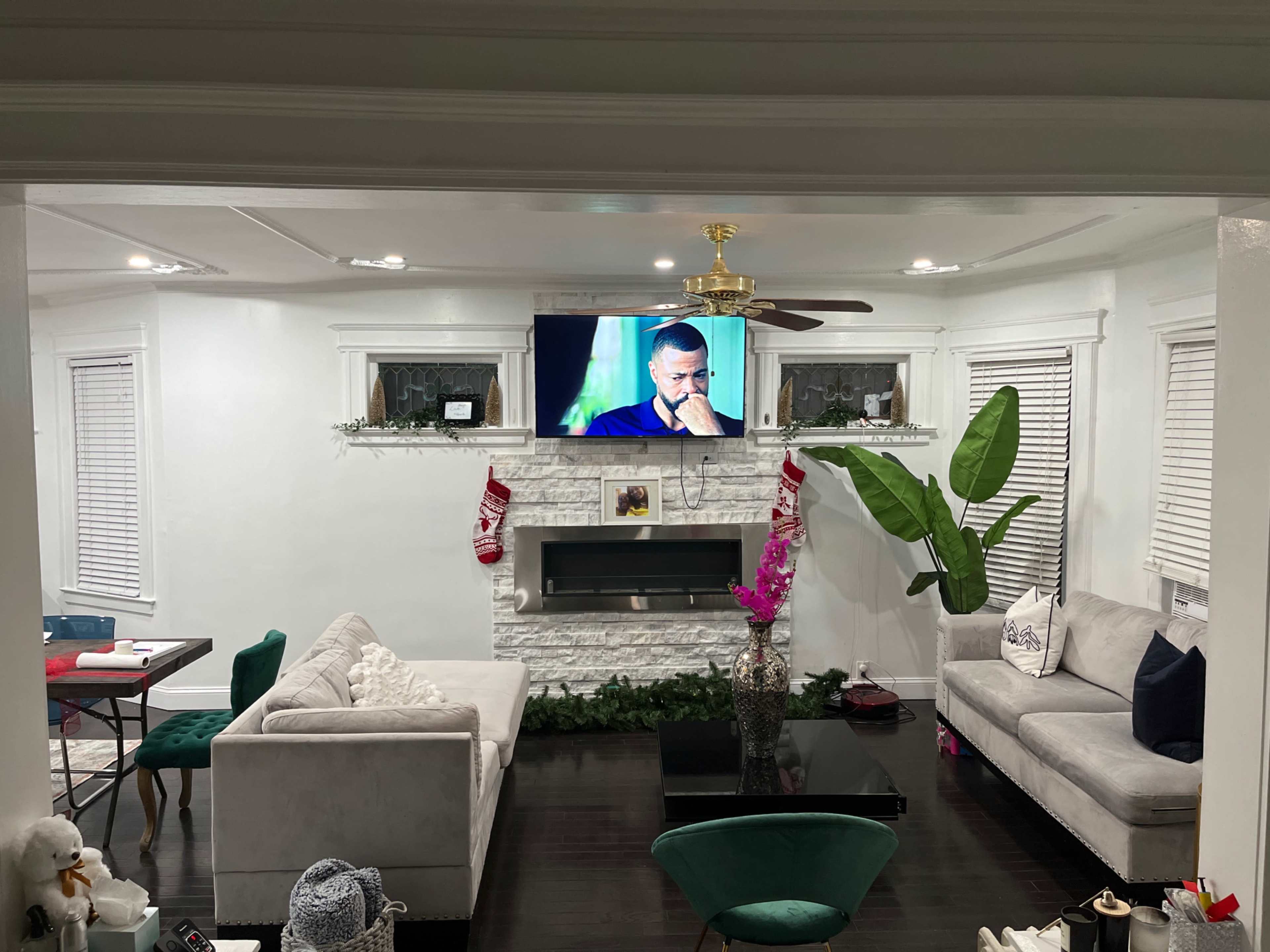The living room features a white marble fireplace, two gray sofas, a large television mounted on the wall, and a green potted plant beside a decorative mantel.