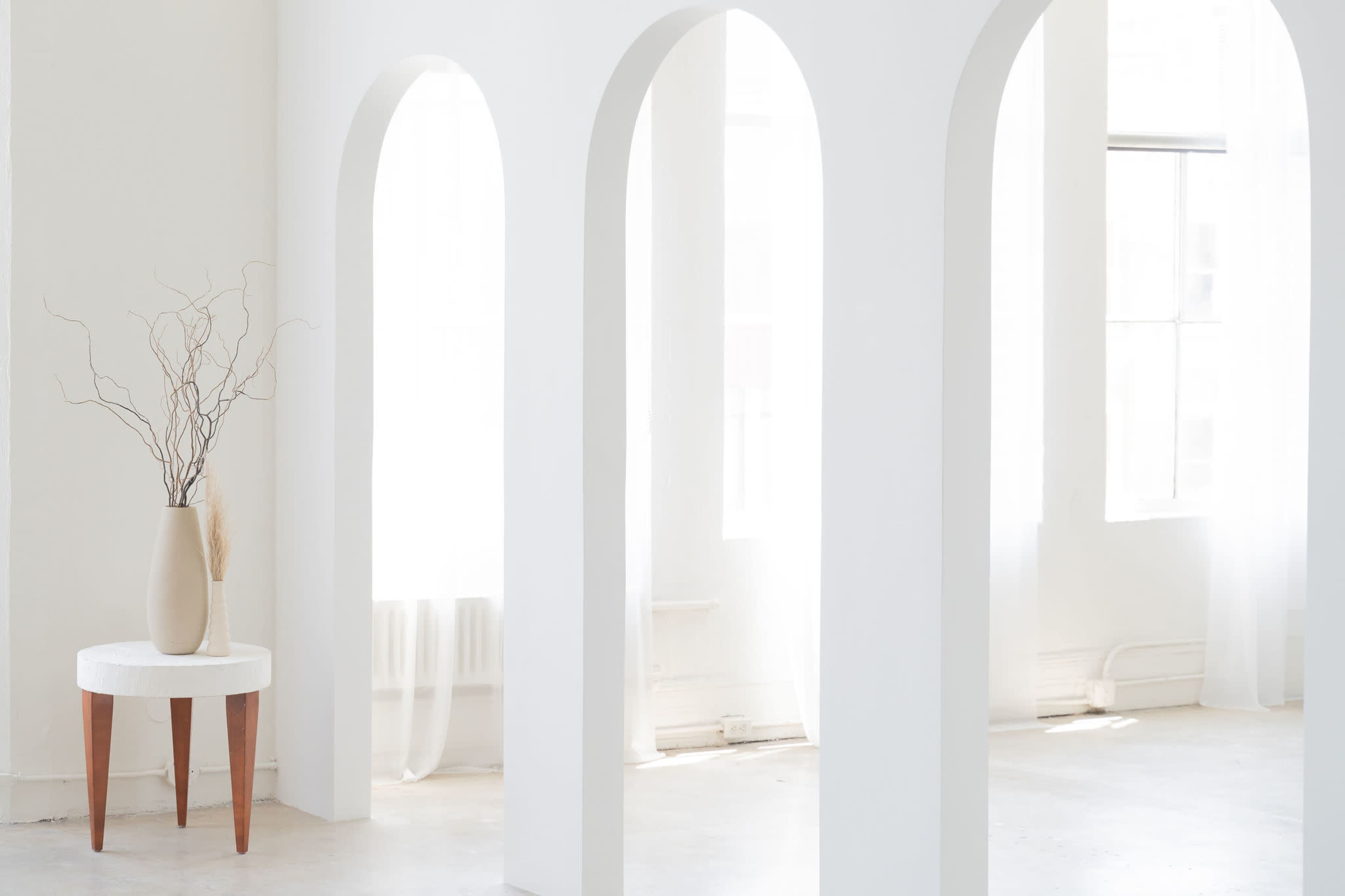 The image displays a minimalist interior with white arches, a round table with a vase containing branches, and large windows allowing natural light.