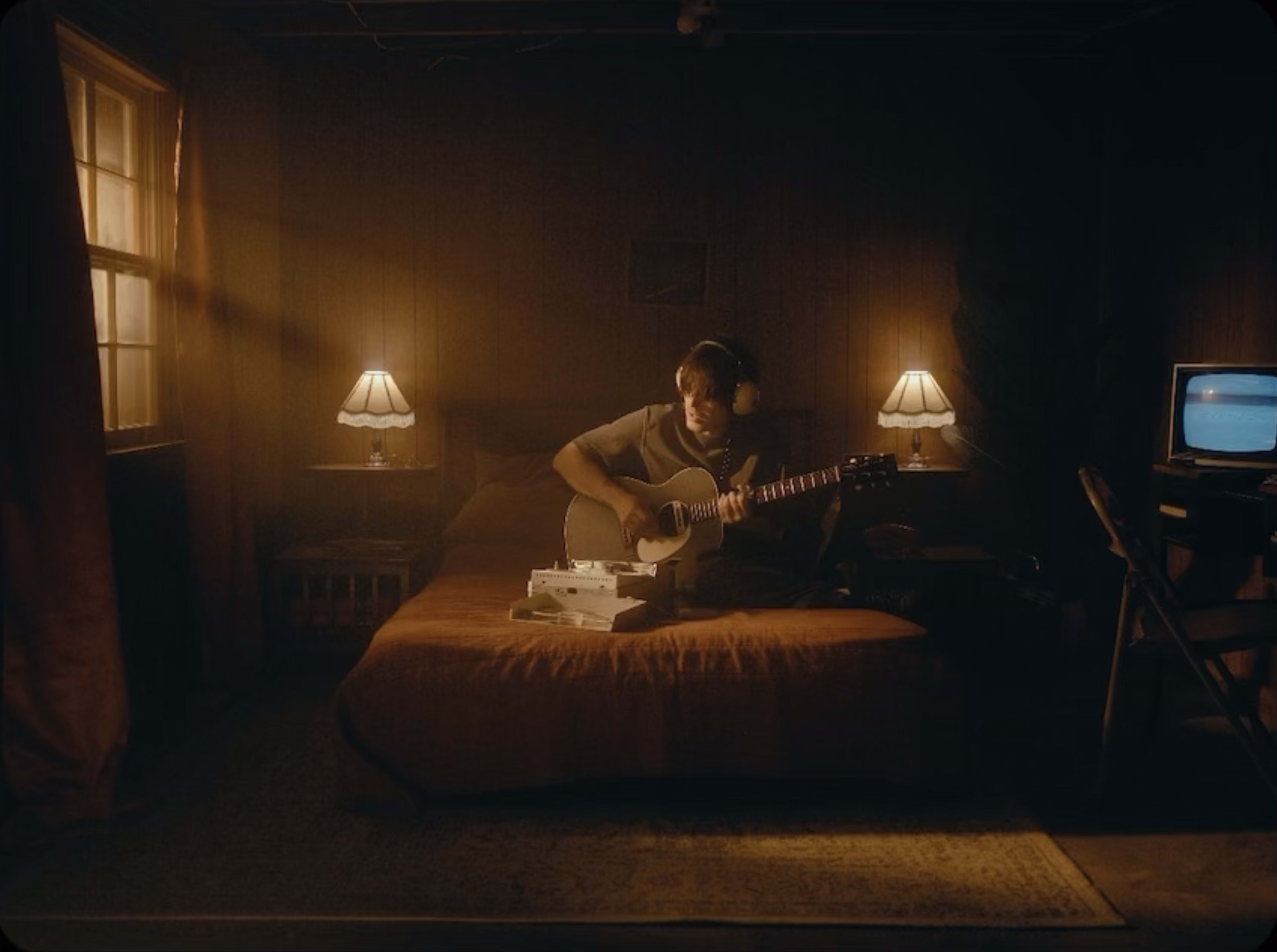 A musician sits on a bed in a dimly lit room, playing an acoustic guitar with a retro television and lamps on either side.