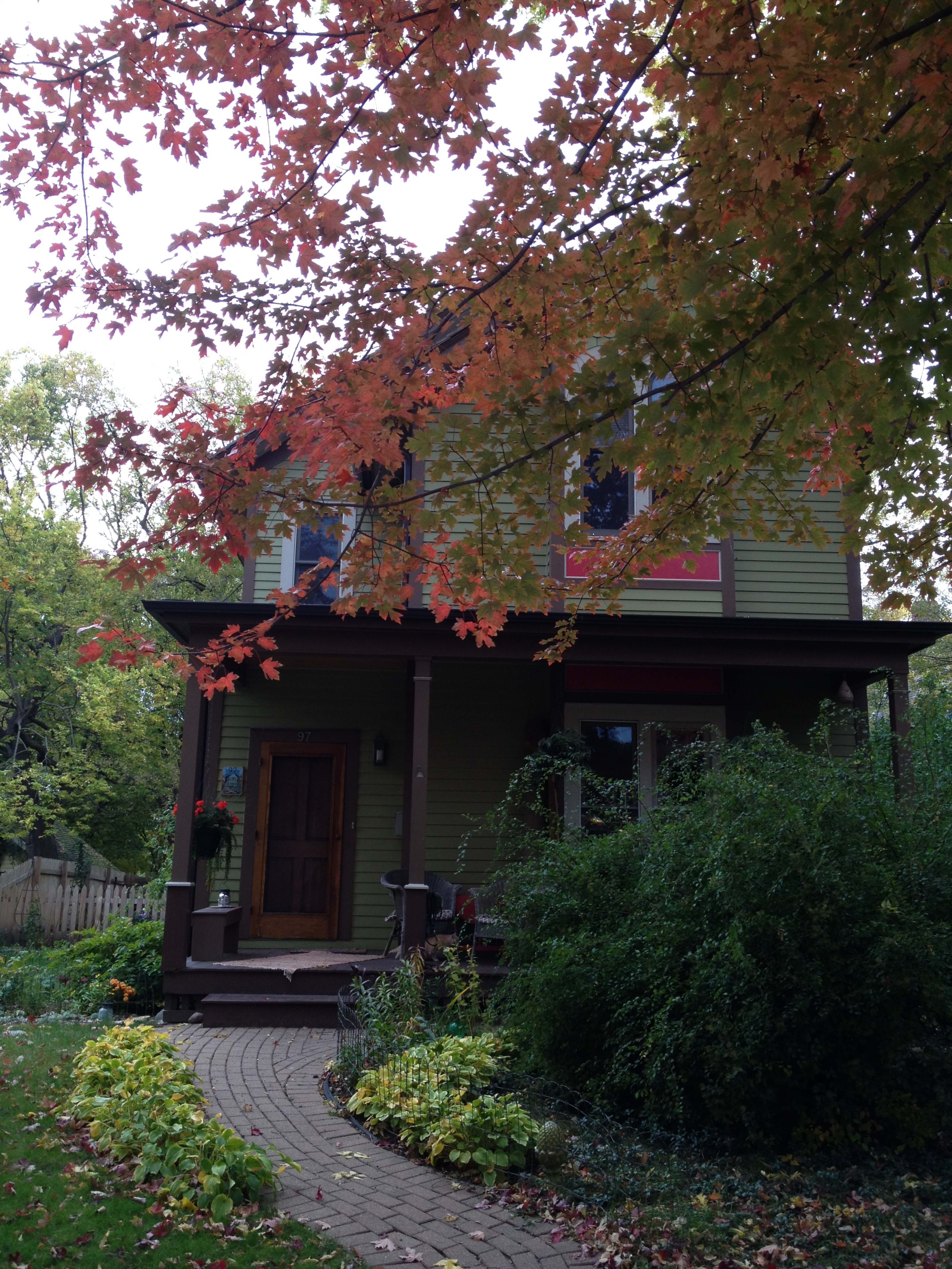 A two-story house with green siding is framed by colorful autumn leaves and a winding brick pathway.