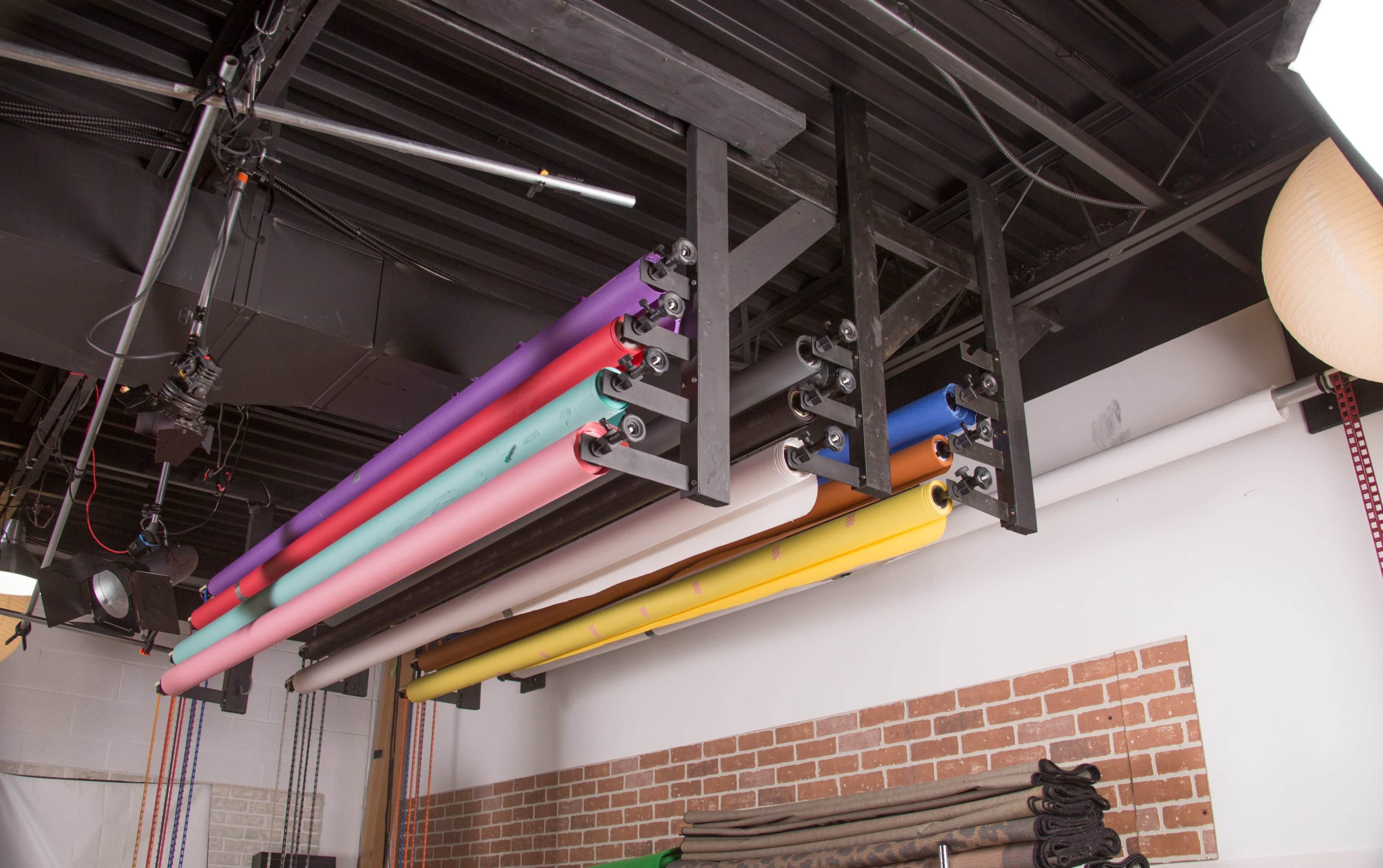 A set of colorful seamless backdrop rolls is mounted on a rack attached to the ceiling in a studio.