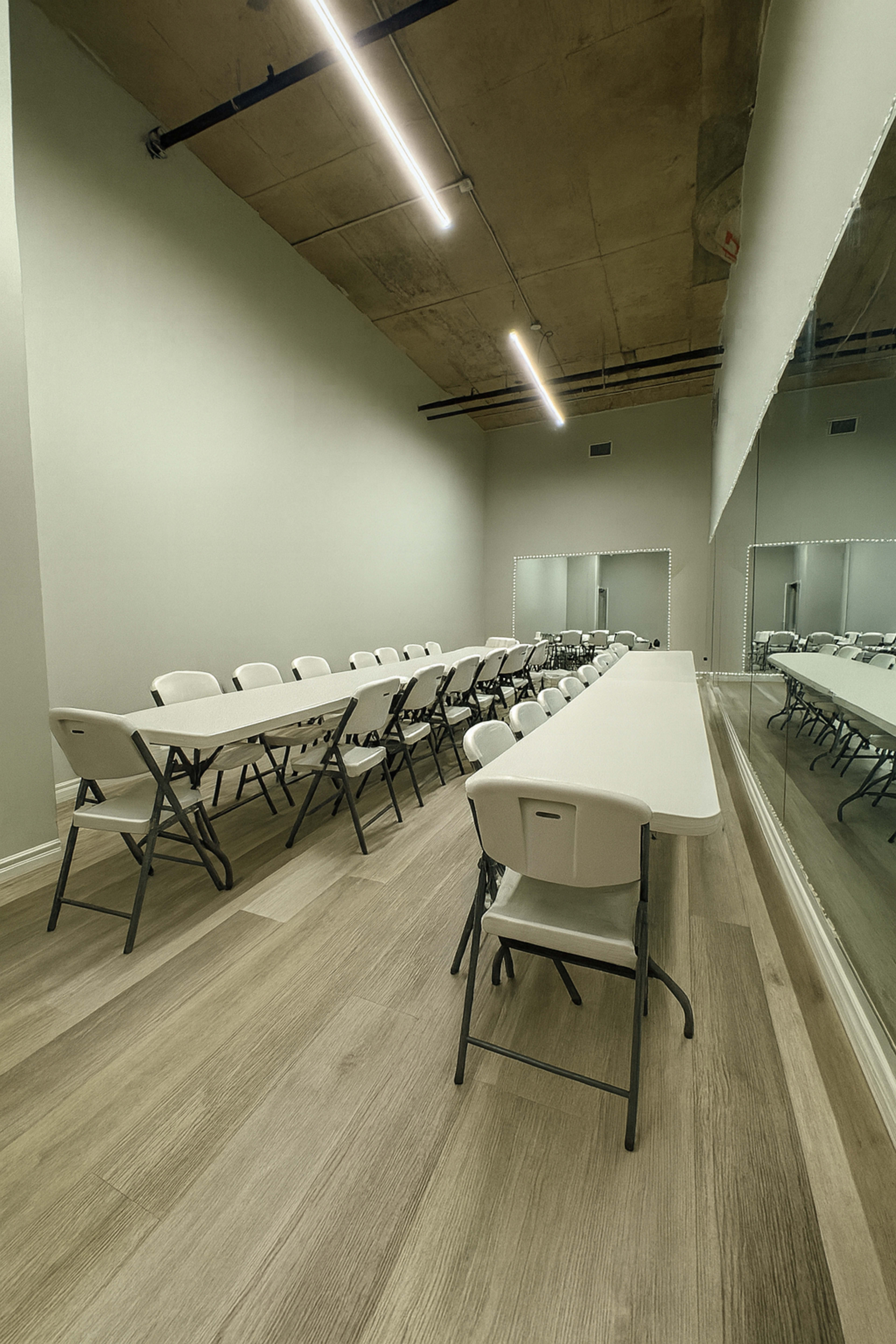 A spacious room features long tables arranged in rows, with white folding chairs positioned around them and mirrors along one wall.