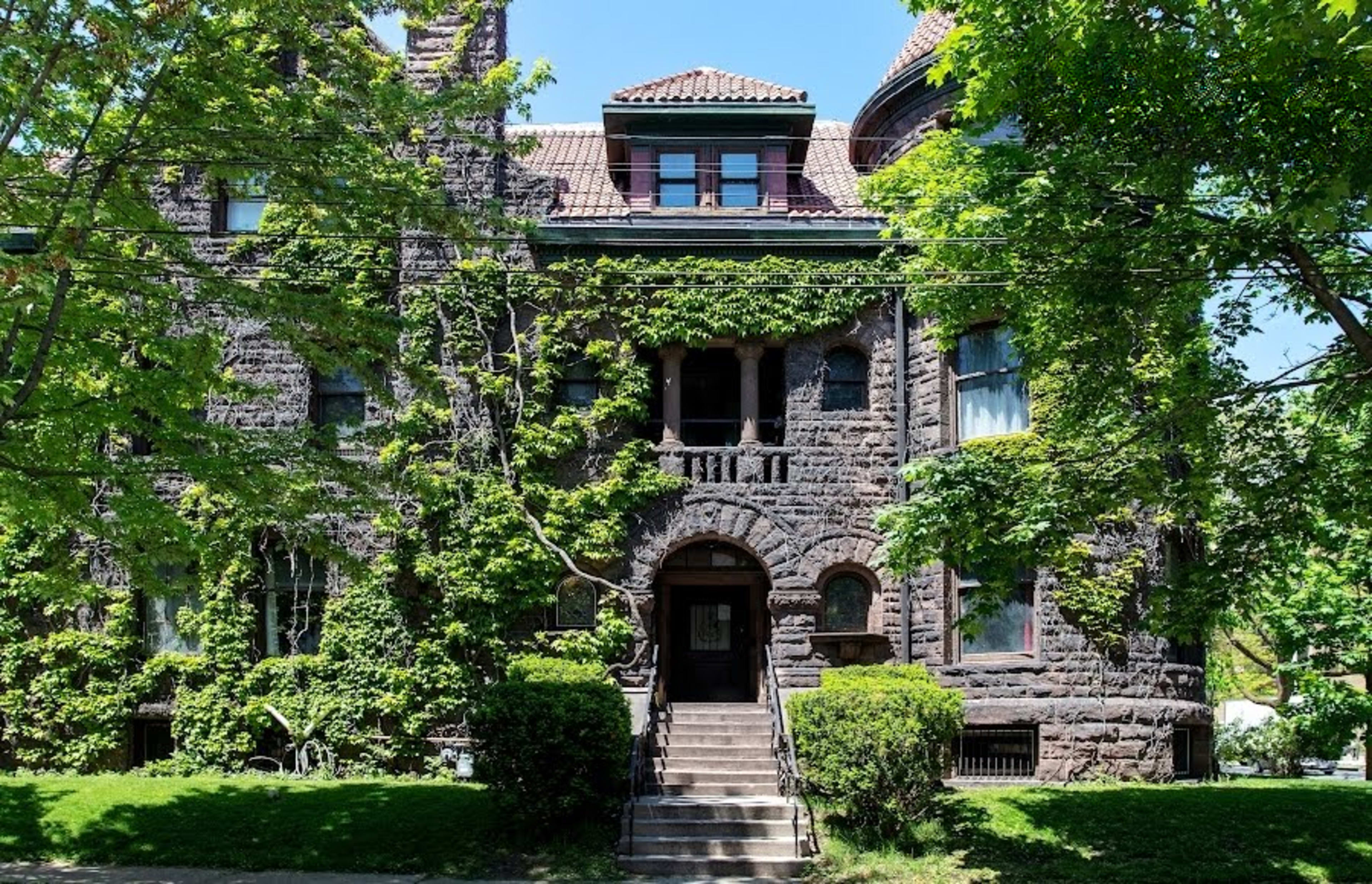 The image shows a historic stone building covered with greenery, featuring large windows and multiple levels.