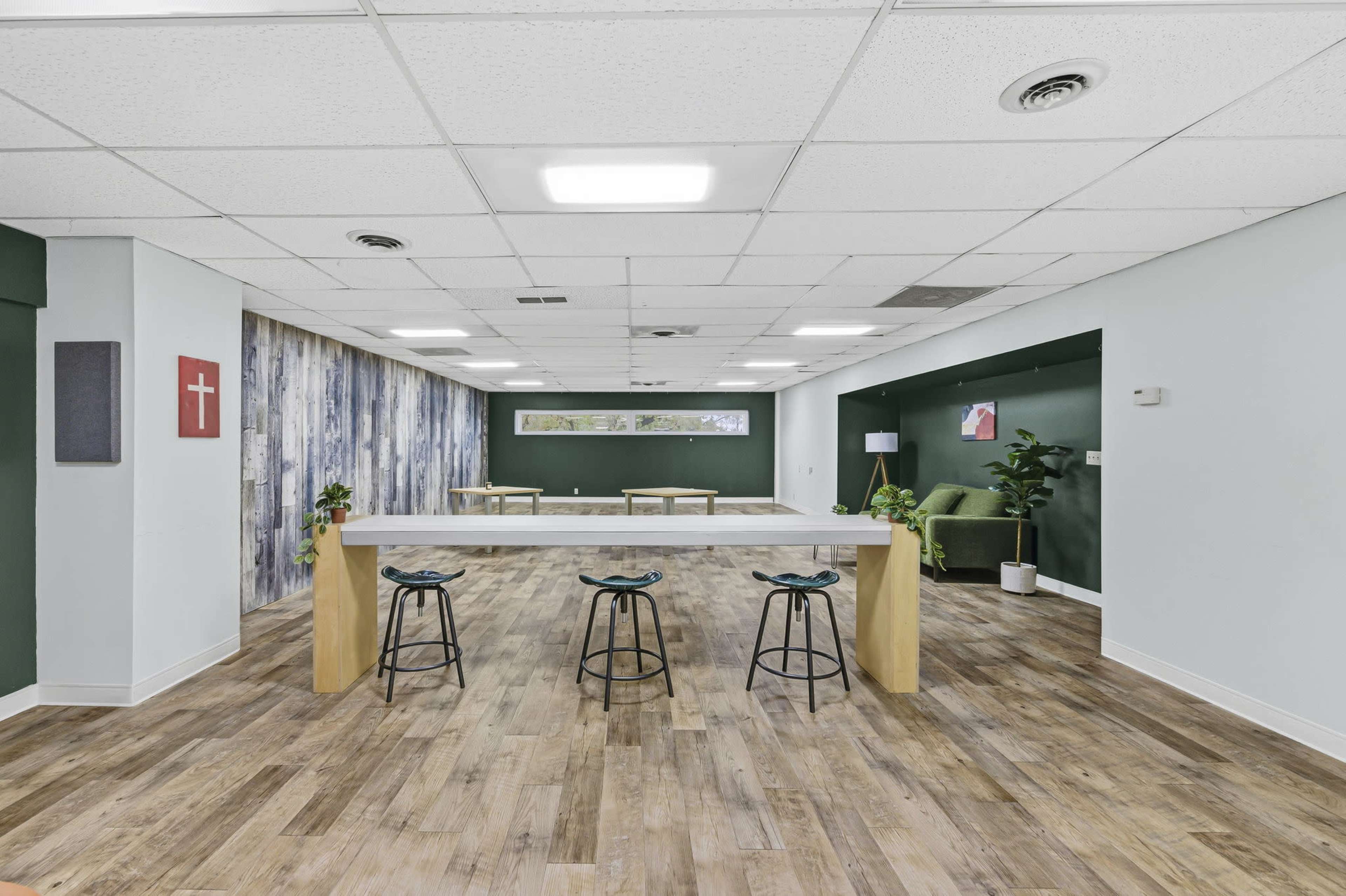 The image shows a modern, spacious communal area with a long table and bar stools, surrounded by green walls and wooden flooring.