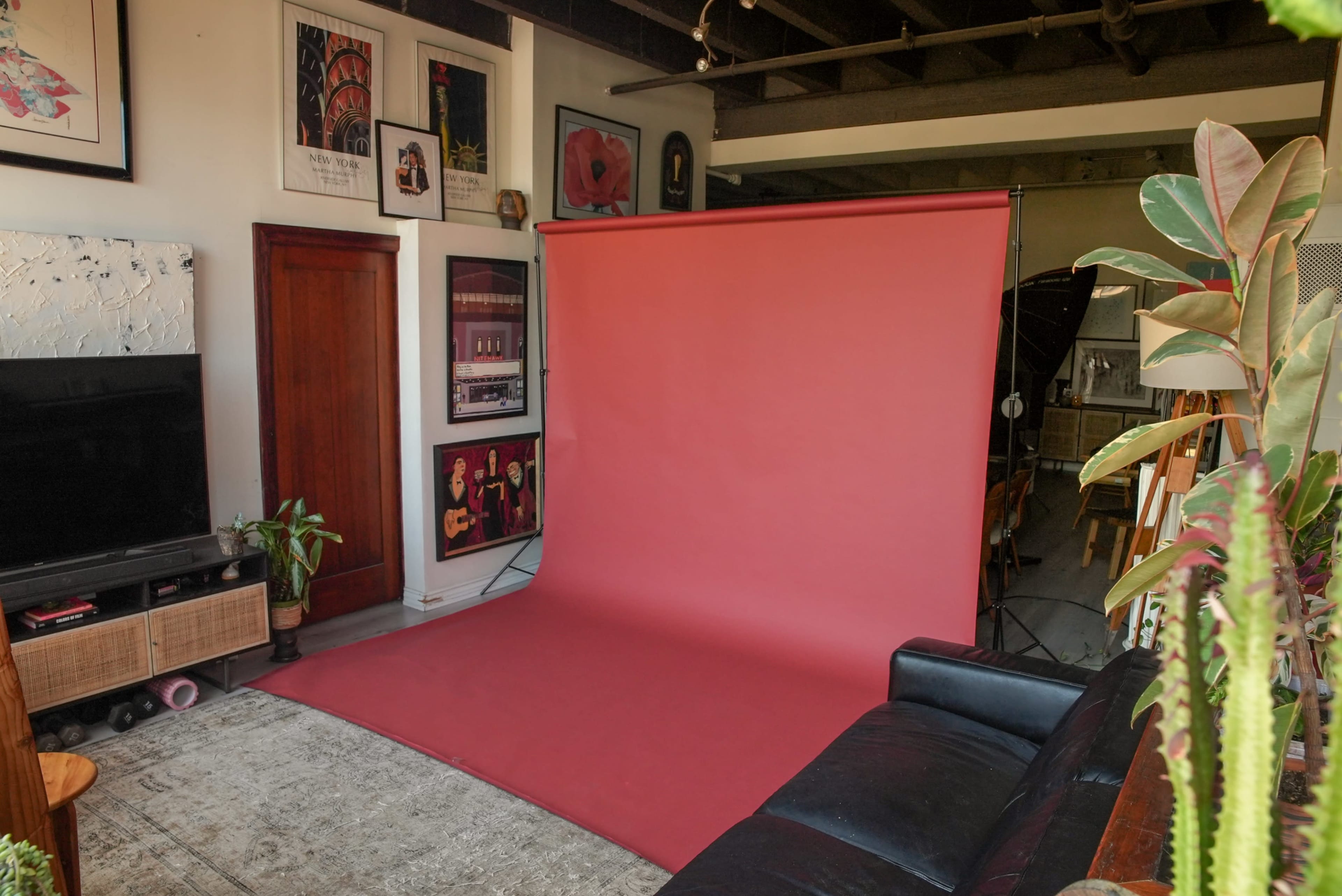 The image shows a spacious room featuring a maroon backdrop setup for photography, a black leather sofa, a patterned rug, and various framed artworks on the walls.