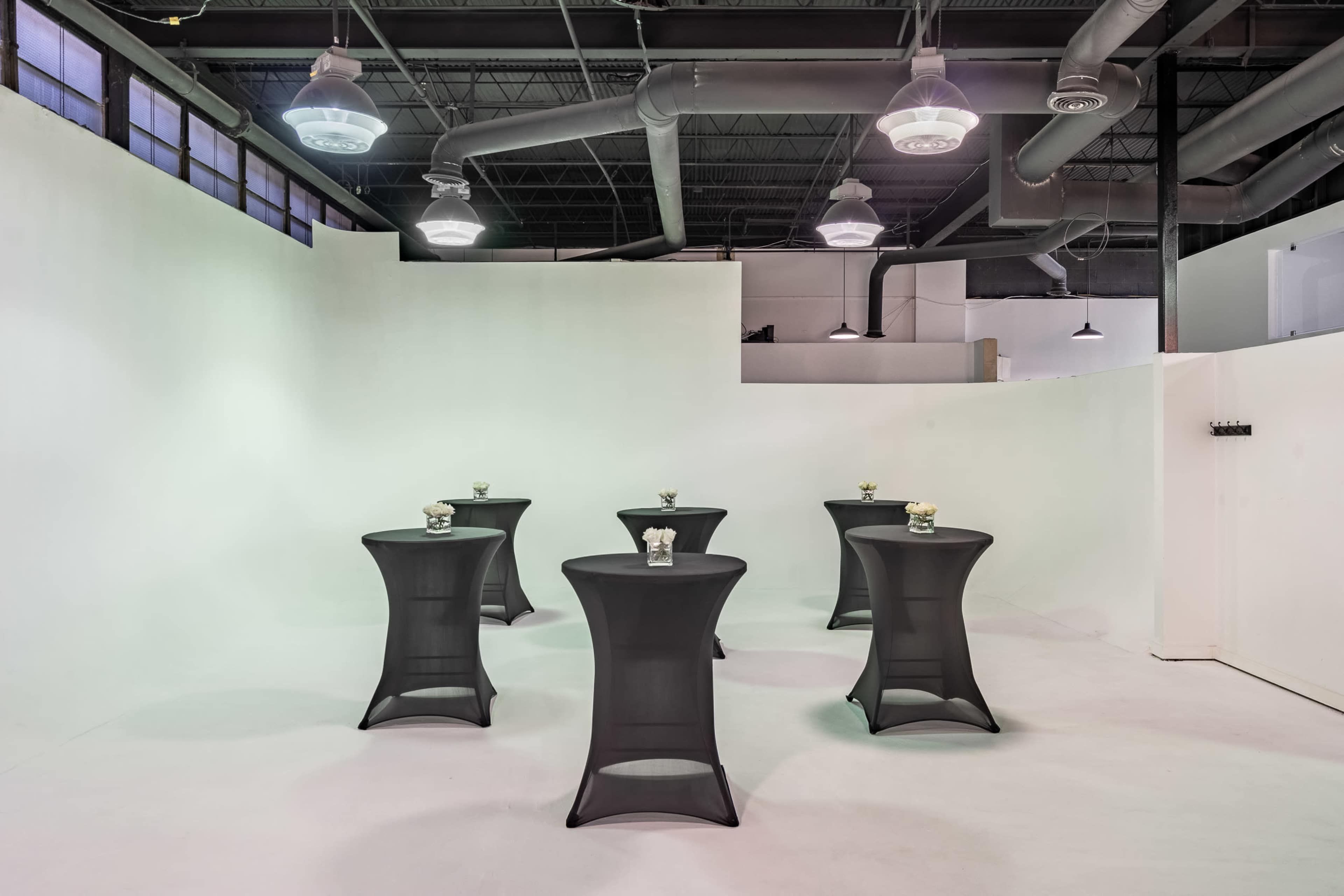 An empty event space with six black cocktail tables set up against a white wall.
