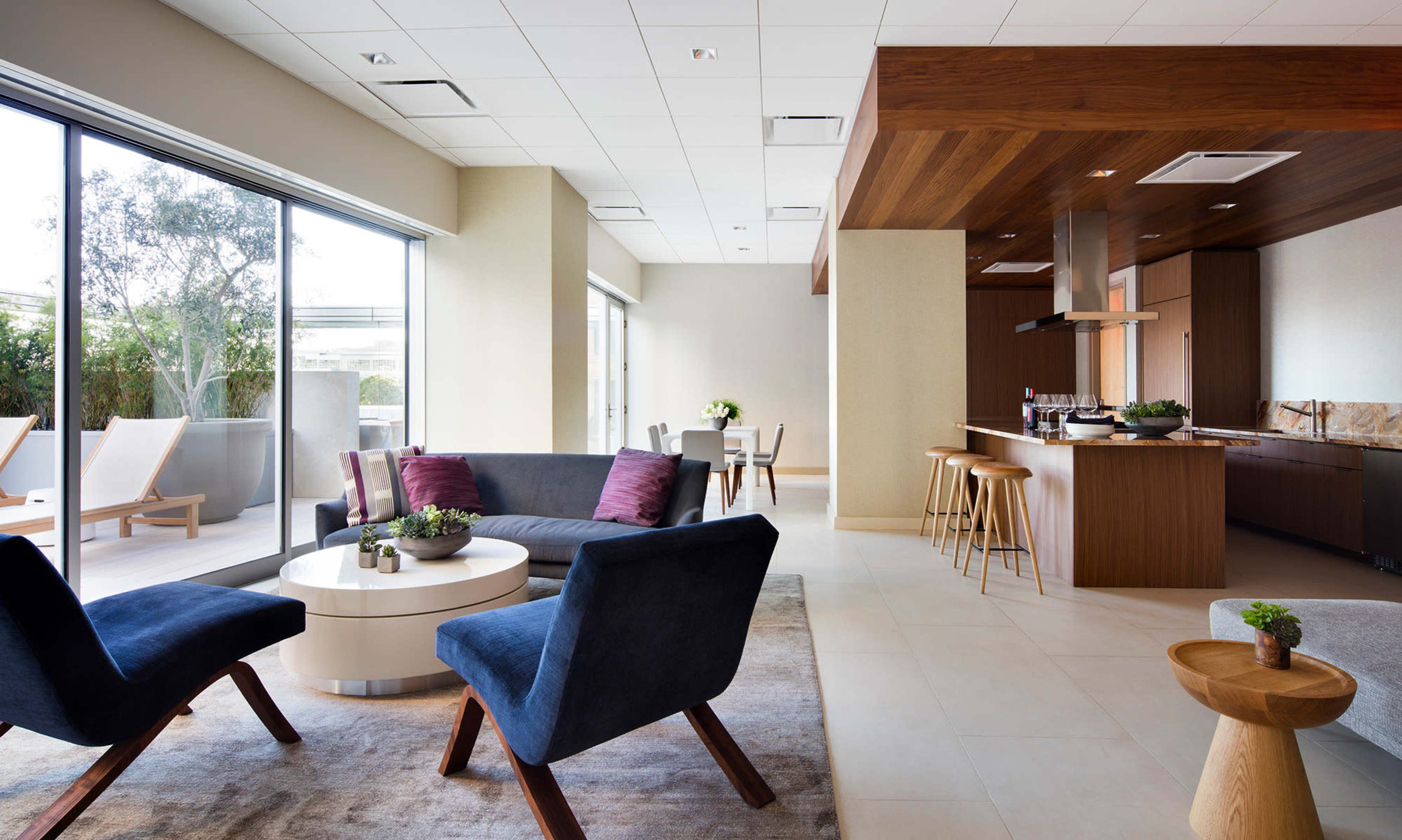 A modern lounge area features seating arrangements with dark blue chairs and a round table, adjacent to a kitchen space and large windows overlooking an outdoor patio.