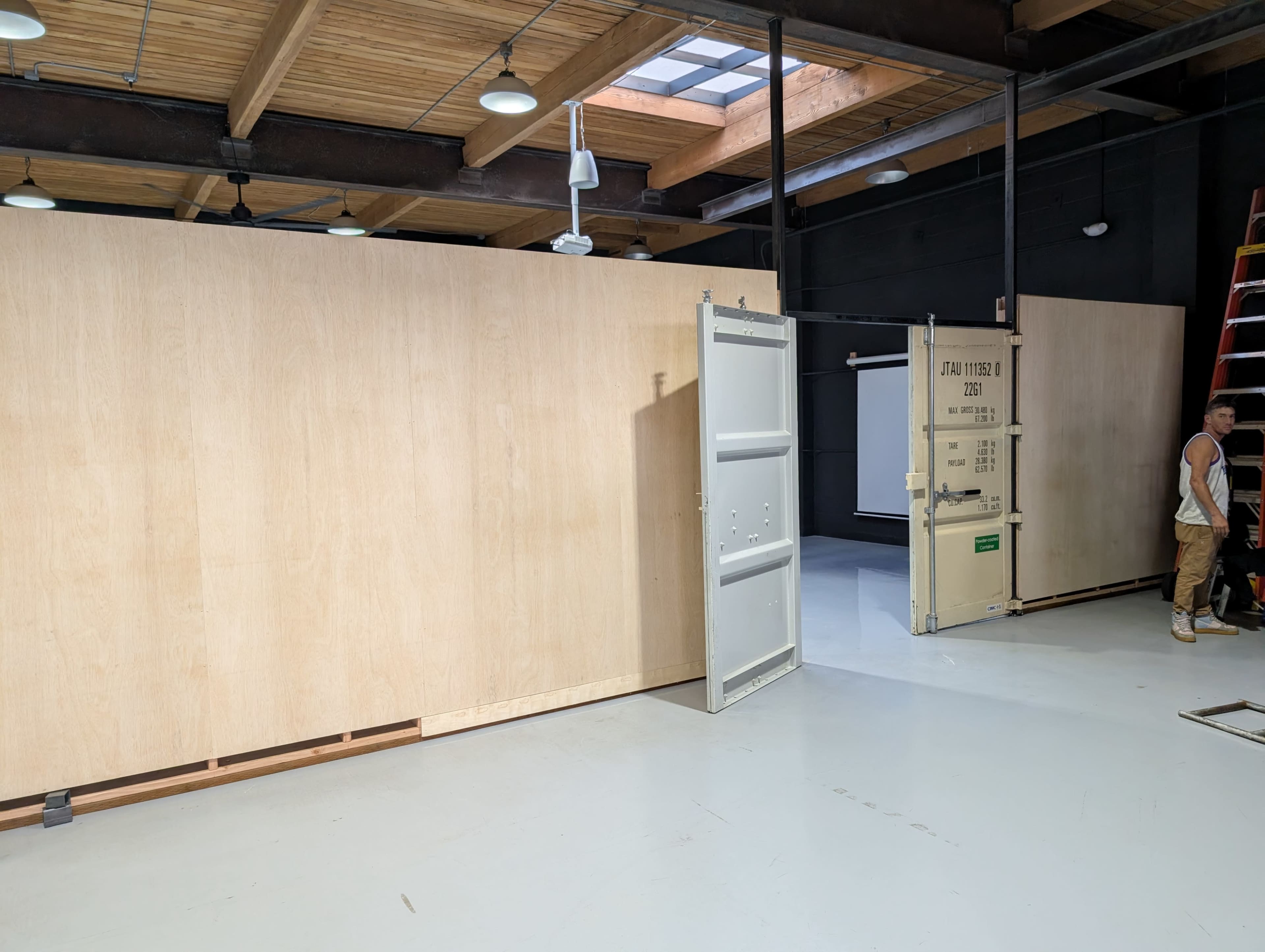 The image shows a spacious interior with plywood walls, an open door leading to another section, and a man standing near a ladder.