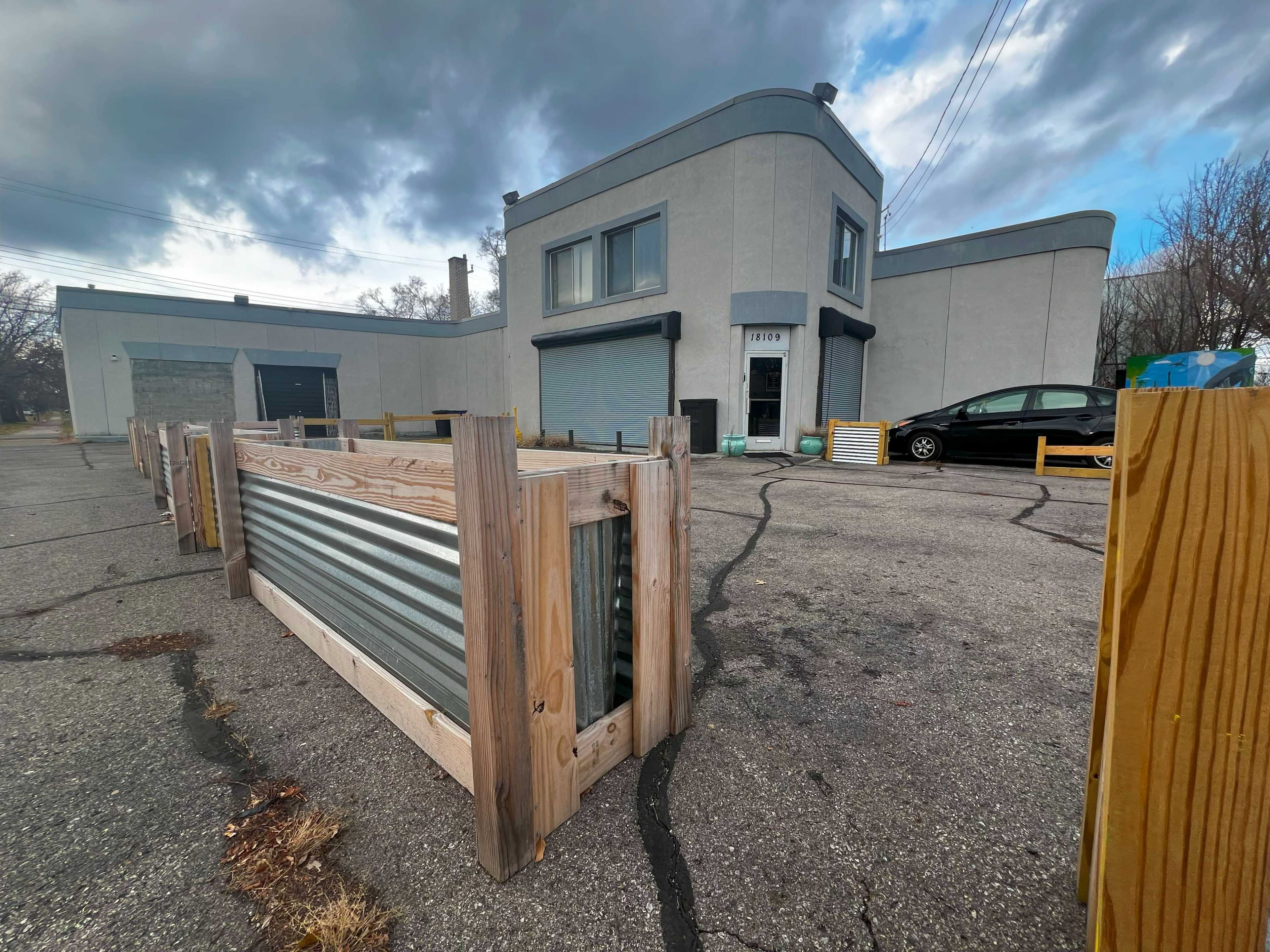 A gray, two-story building with a curved roof features a closed entrance, surrounded by a fenced parking lot with metal barriers.