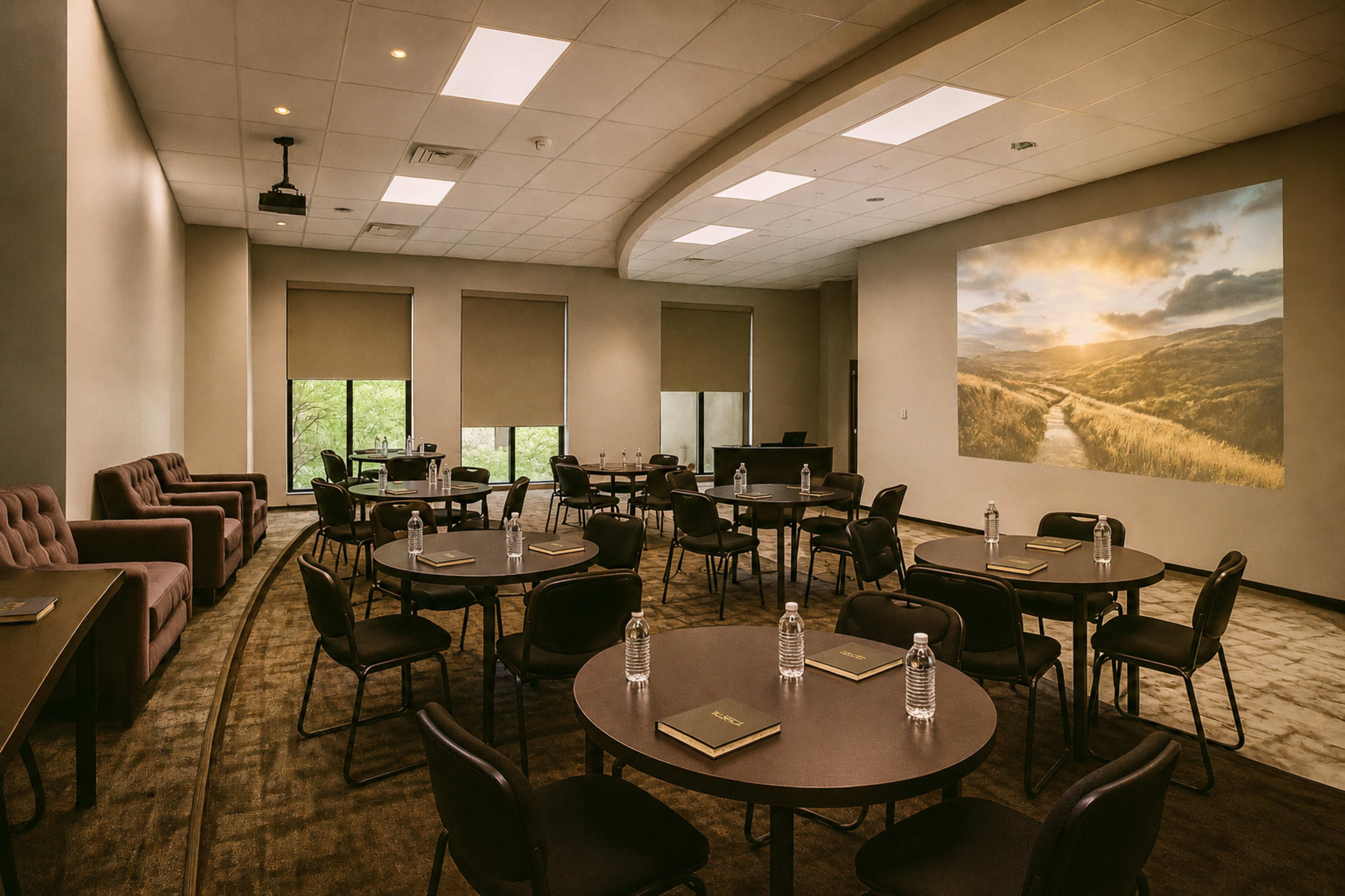 The image shows a conference room with several round tables and chairs arranged neatly, featuring large windows and a mural of a scenic landscape on one wall.