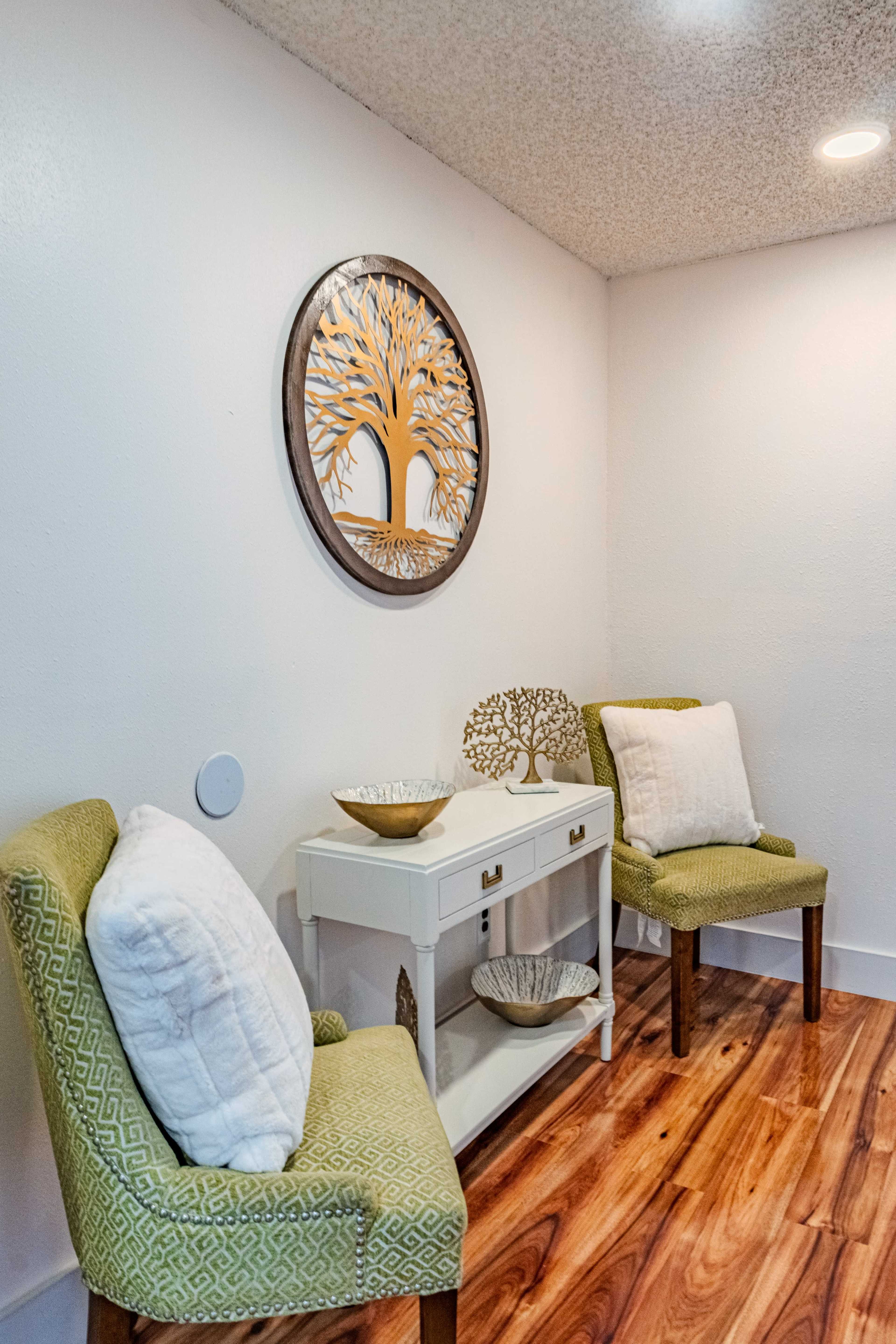 The image shows a small, well-lit entryway with two green chairs, a white console table, and decorative items, including a round wall art featuring a tree design.