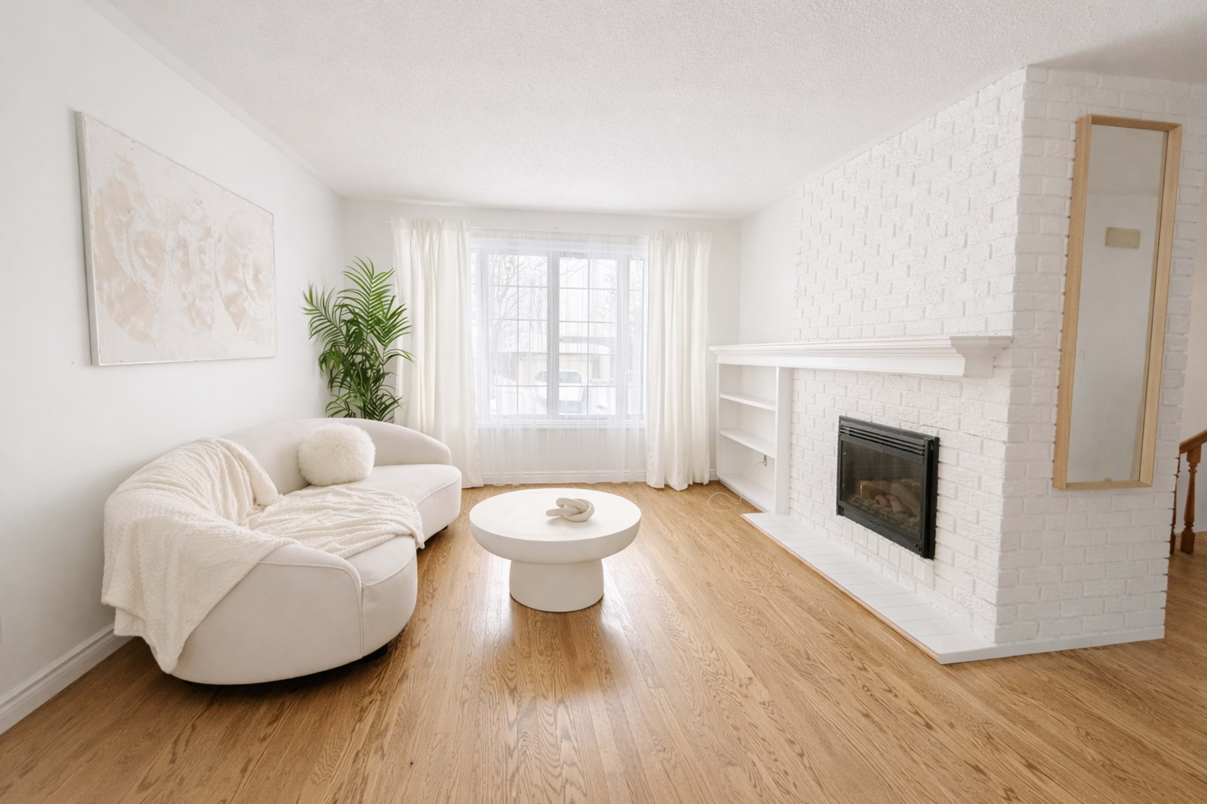 The scene features a modern living room with a white sectional sofa, a round coffee table, a fireplace set in a white brick wall, and large windows allowing natural light to fill the space.